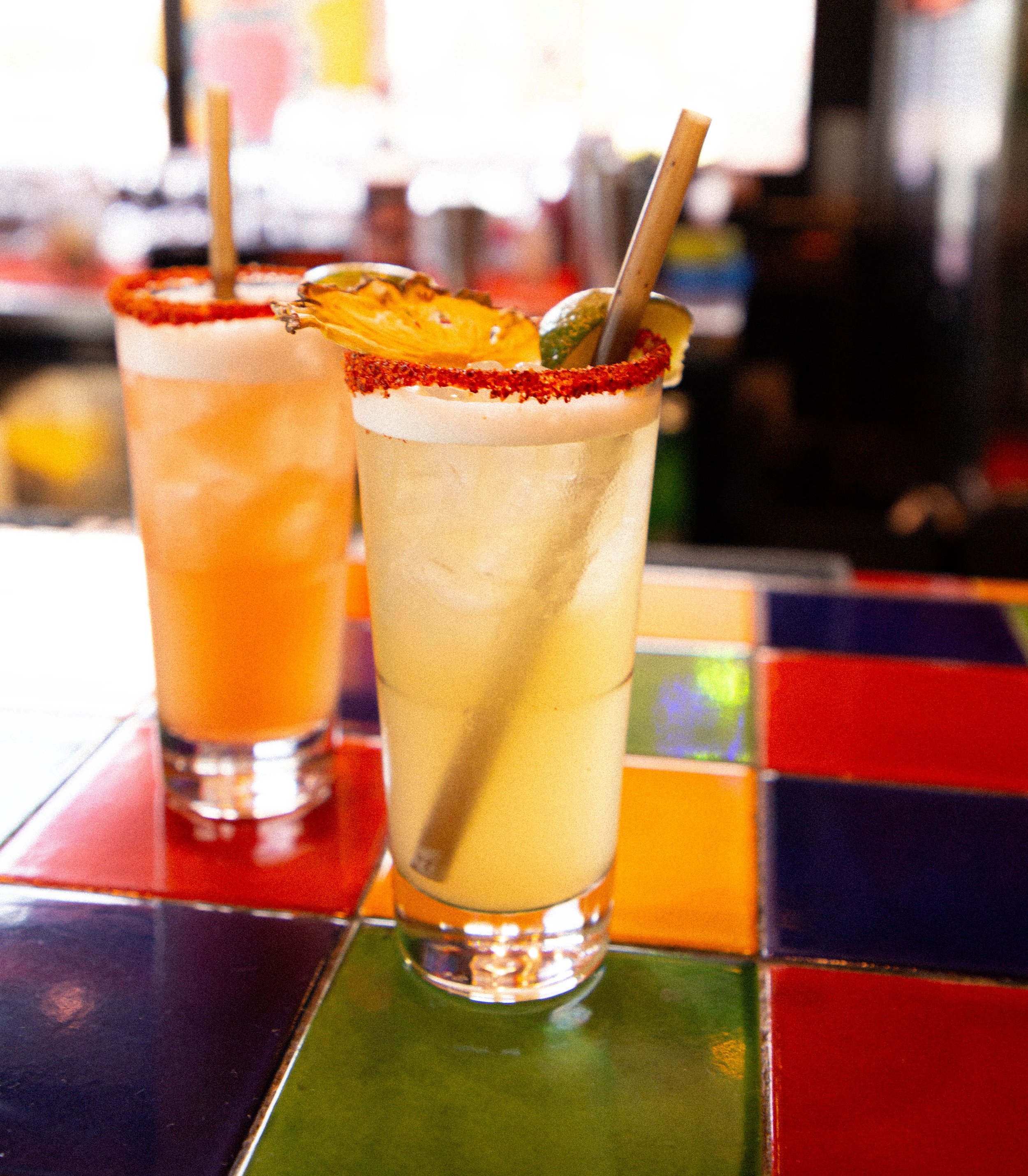Two margarita cocktails on a table at a colorful restaurant or bar. The drinks have salt-rimmed glasses, garnished with lime, served with straws.
