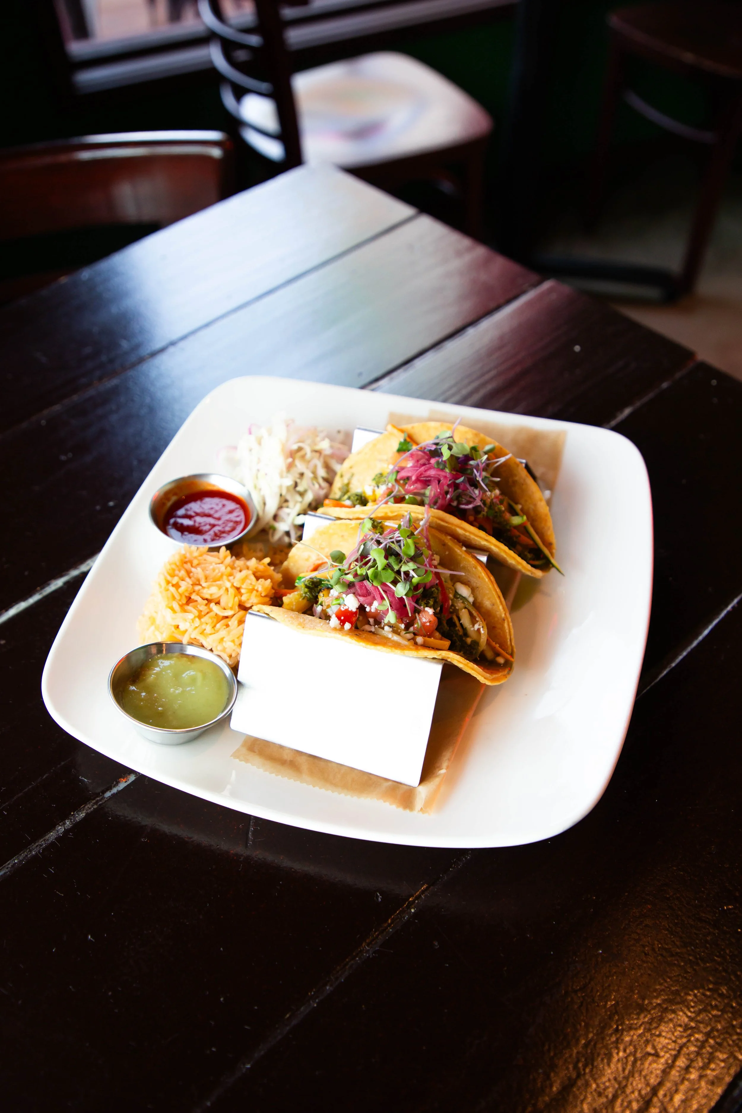 A plate of Mexican tacos with microgreens on top, served with sides of rice, coleslaw, and two types of salsa in small metal cups, placed on a white square plate.