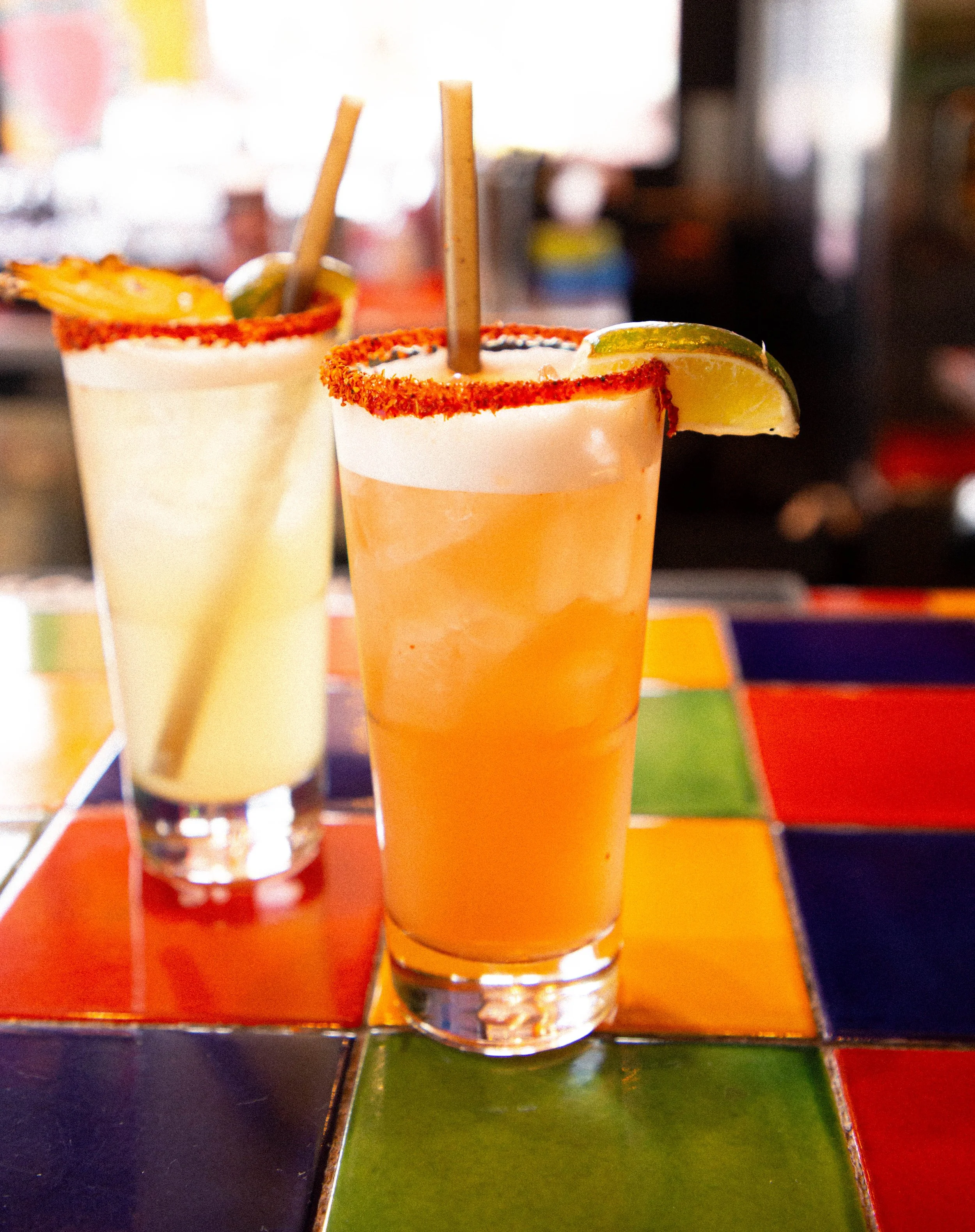 Close-up of two margarita cocktails with salted rims, lime wedges, and straws, on a colorful tiled table in a restaurant or bar setting.