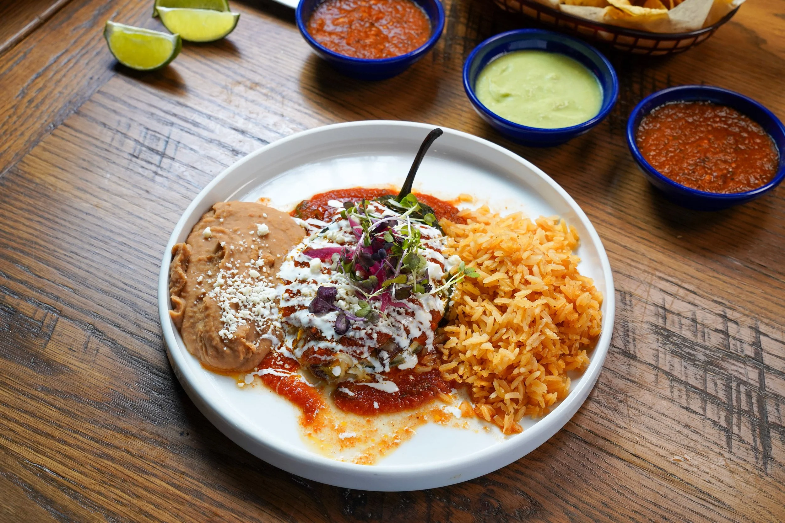 Plate of Mexican food with rice, enchiladas topped with white sauce and garnished with microgreens, served with three bowls of salsa and guacamole and lime wedges on a wooden table.