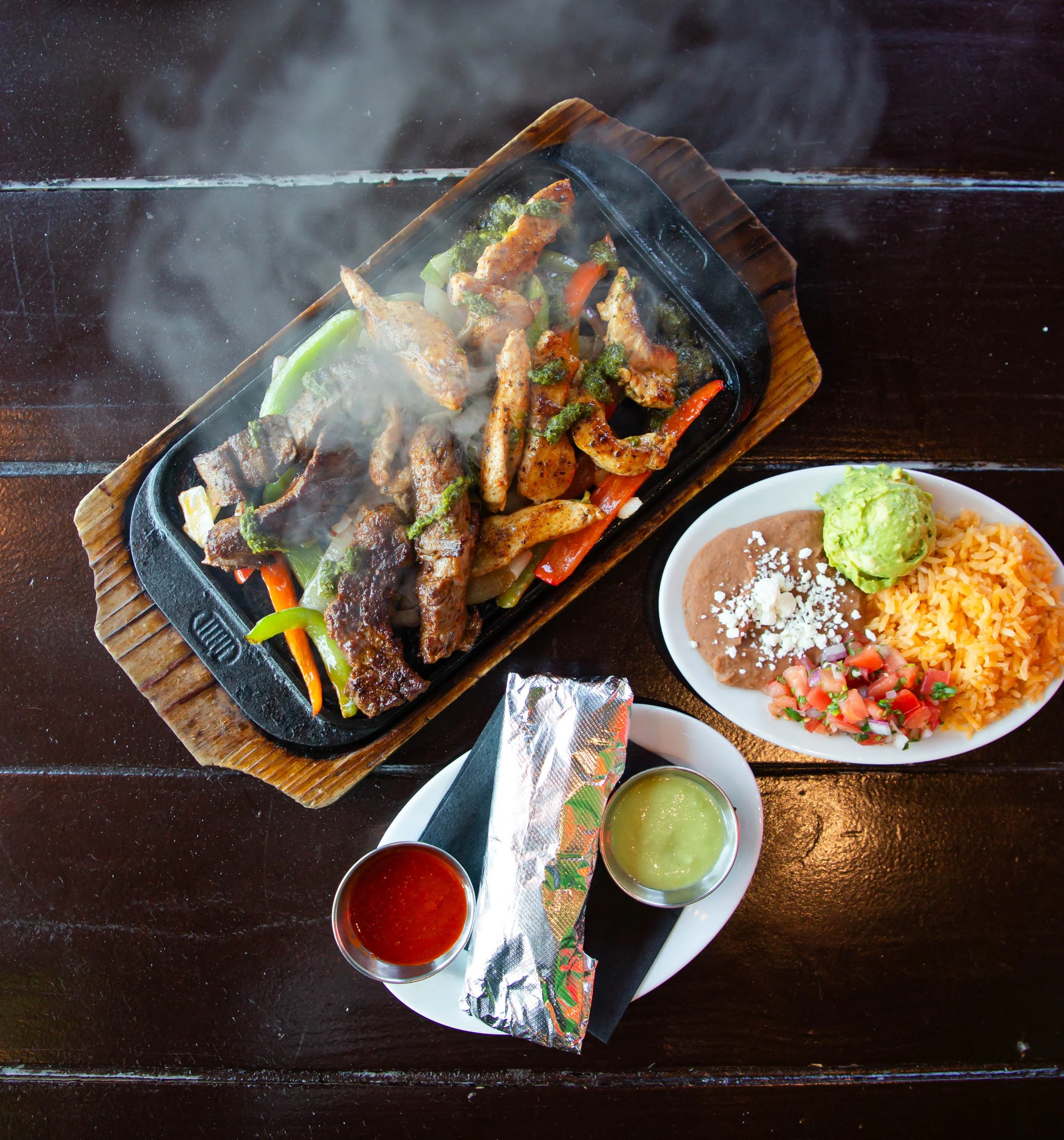 A sizzling fajita dish with grilled chicken, sautéed vegetables including bell peppers and broccoli, served on a cast iron skillet on a wooden tray, with bowls of salsa, guacamole, rice, and beans on a dark wooden table.