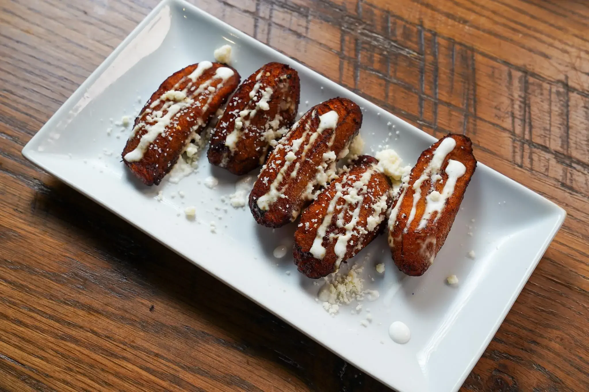 Four fried plantains drizzled with white sauce and garnished with crumbled cheese, served on a white rectangular plate on a wooden table.
