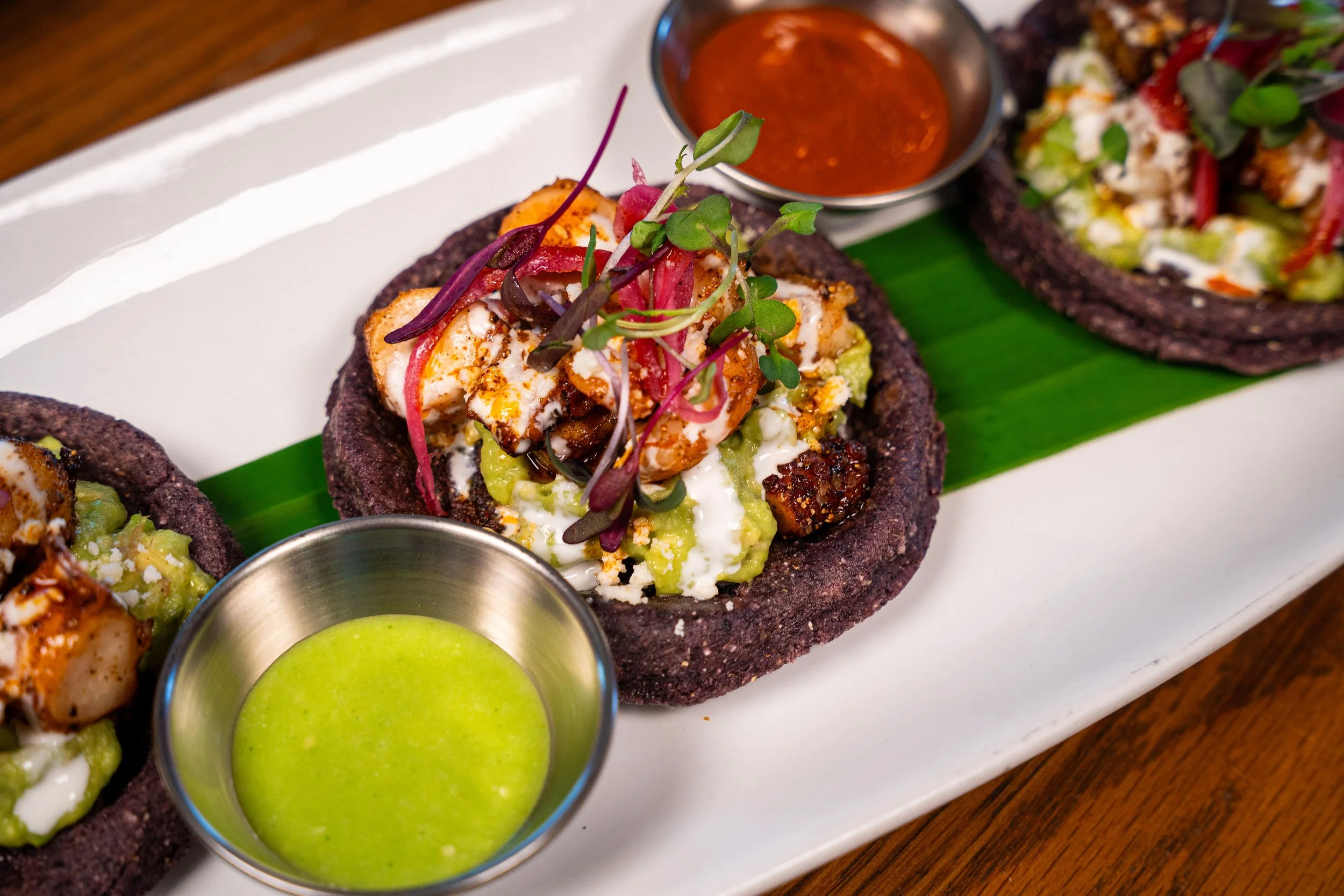 Close-up of a plate with black charcoal taco shells filled with grilled chicken, salsa, and microgreens, serving with a side of green sauce and red sauce.