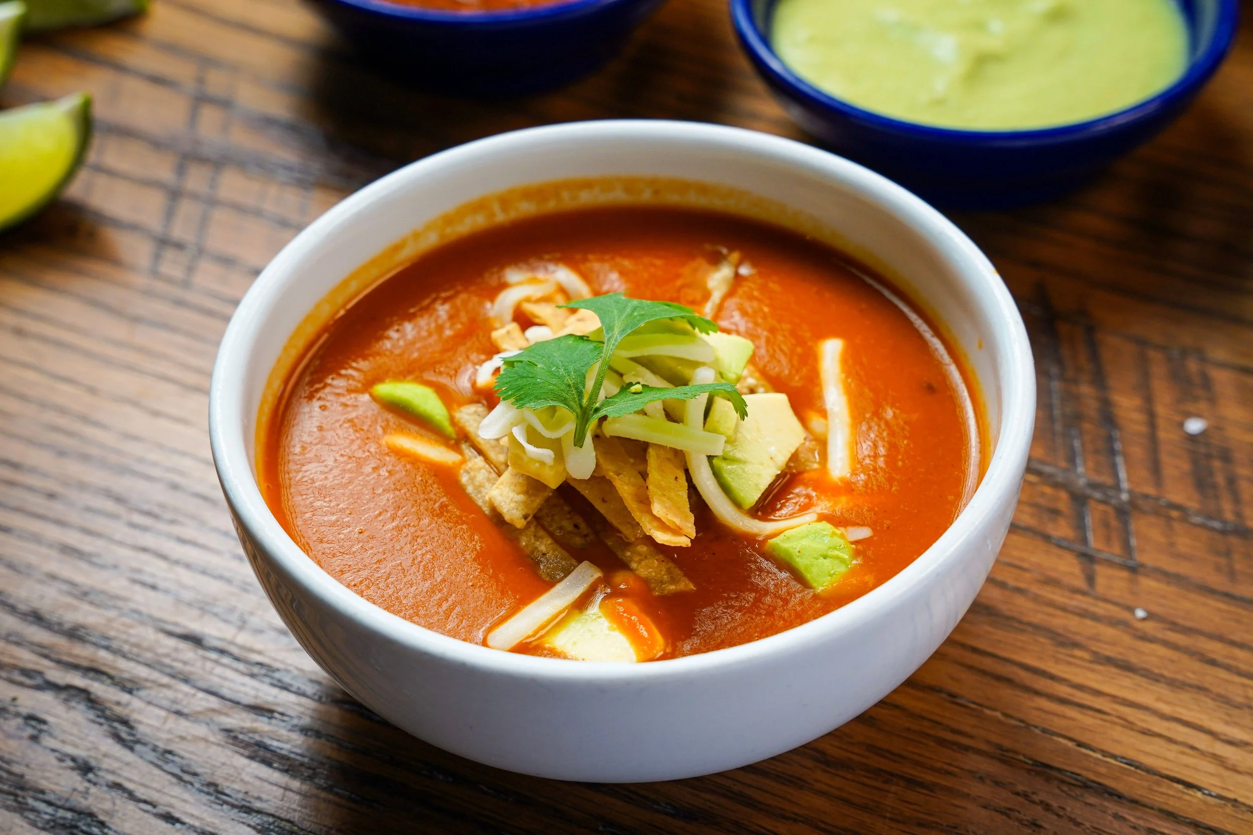 A bowl of red tomato-based soup garnished with sliced avocado, shredded cheese, and a sprig of cilantro, placed on a wooden table with two bowls of green guacamole and a lime wedge partially visible.