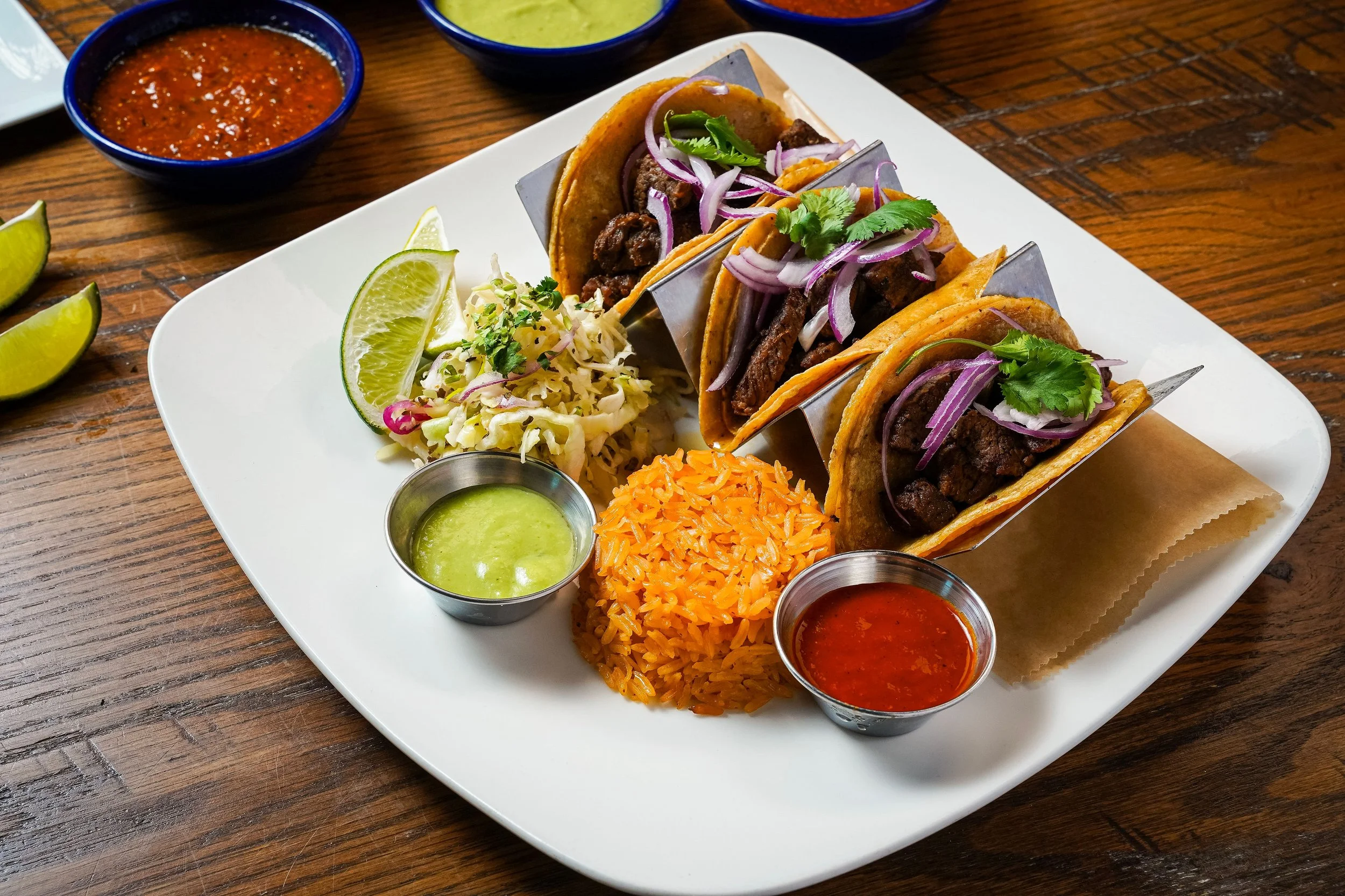 Three beef tacos with onions, cilantro, and sauce on a white plate, accompanied by lime wedges, shredded cabbage, rice, and salsa, on a wooden table.