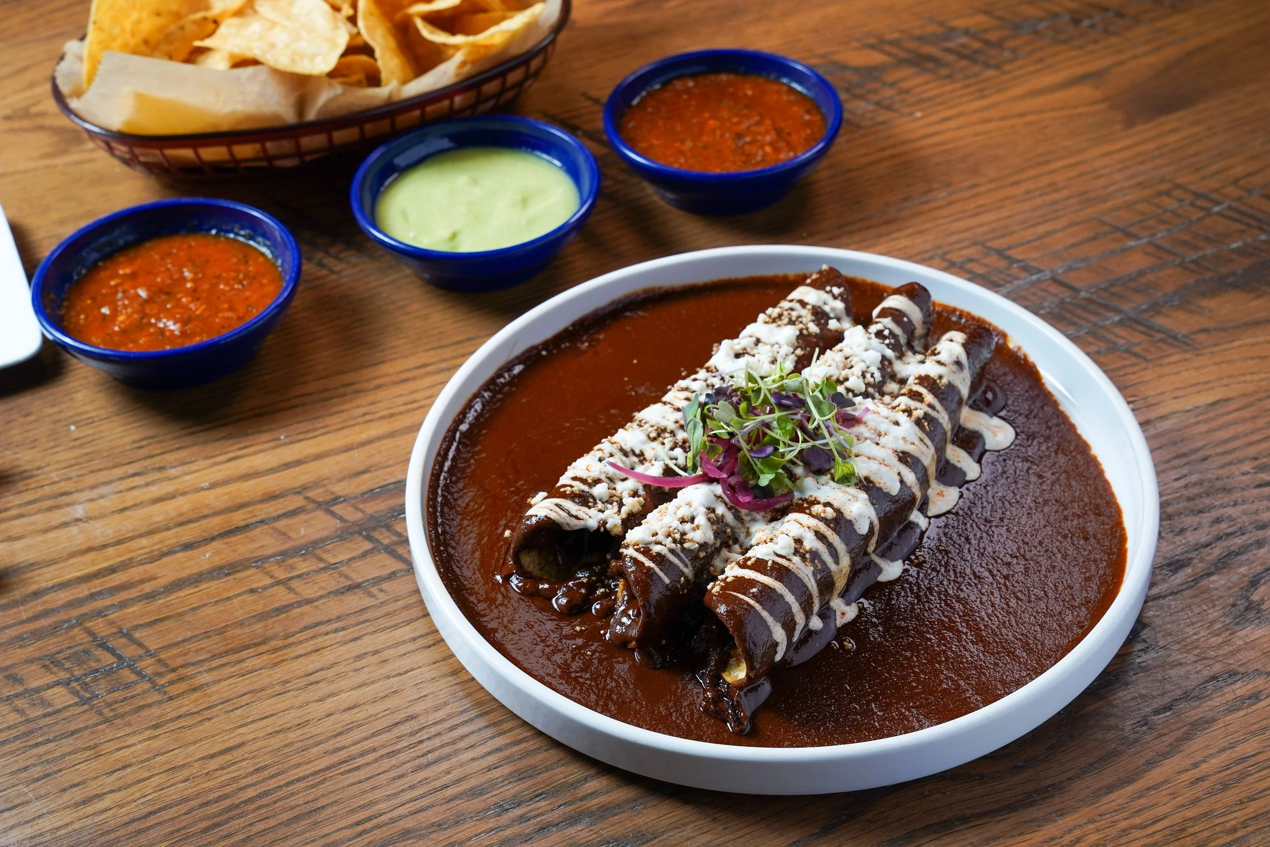 A plate of Mexican enchiladas topped with sauce, cheese, and microgreens, accompanied by small bowls of salsa and guacamole, with a basket of tortilla chips in the background on a wooden table.