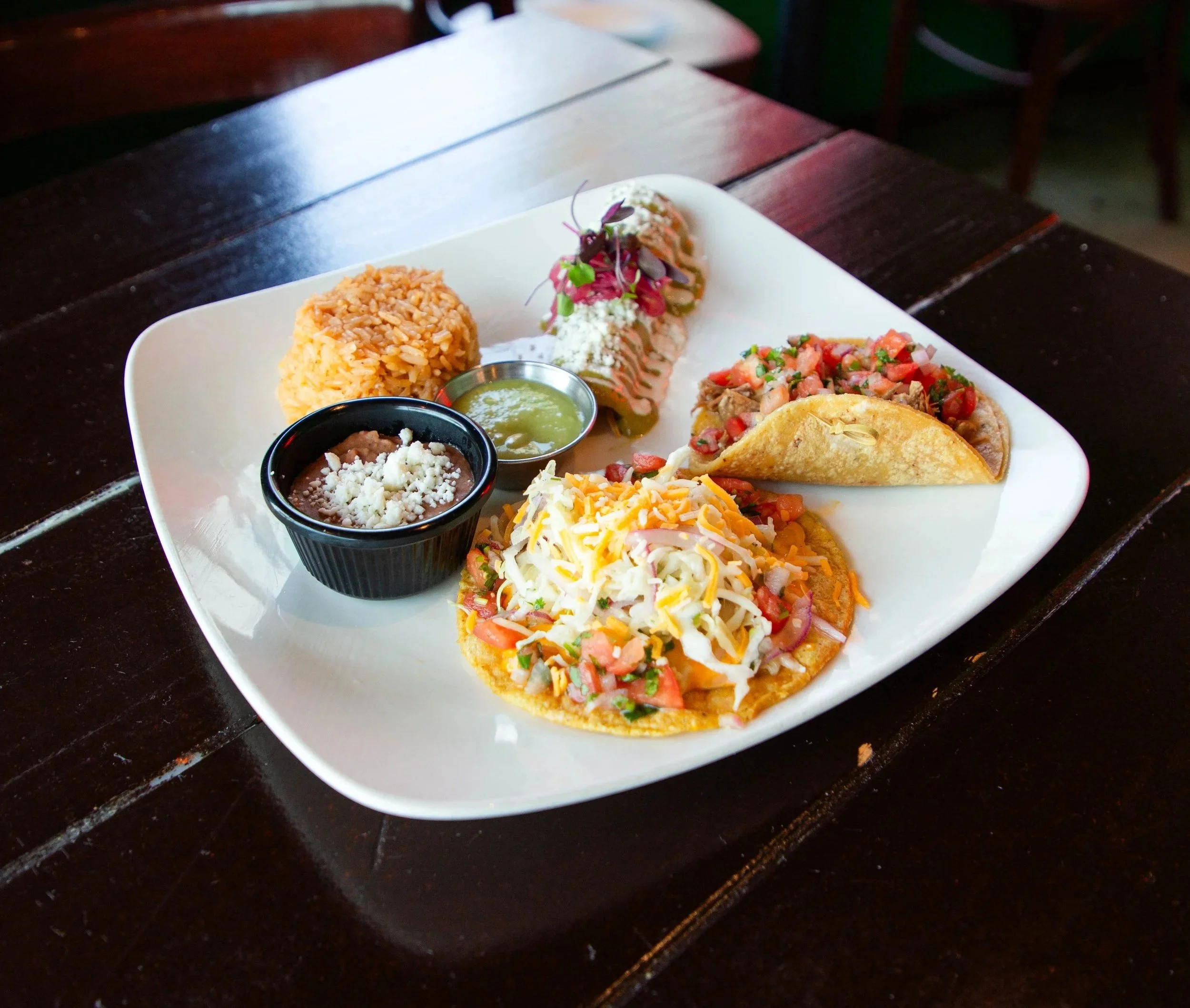 Plate of Mexican food including rice, grilled meat tacos with salsa and cheese, refried beans with cheese, and small containers of green and brown sauces.