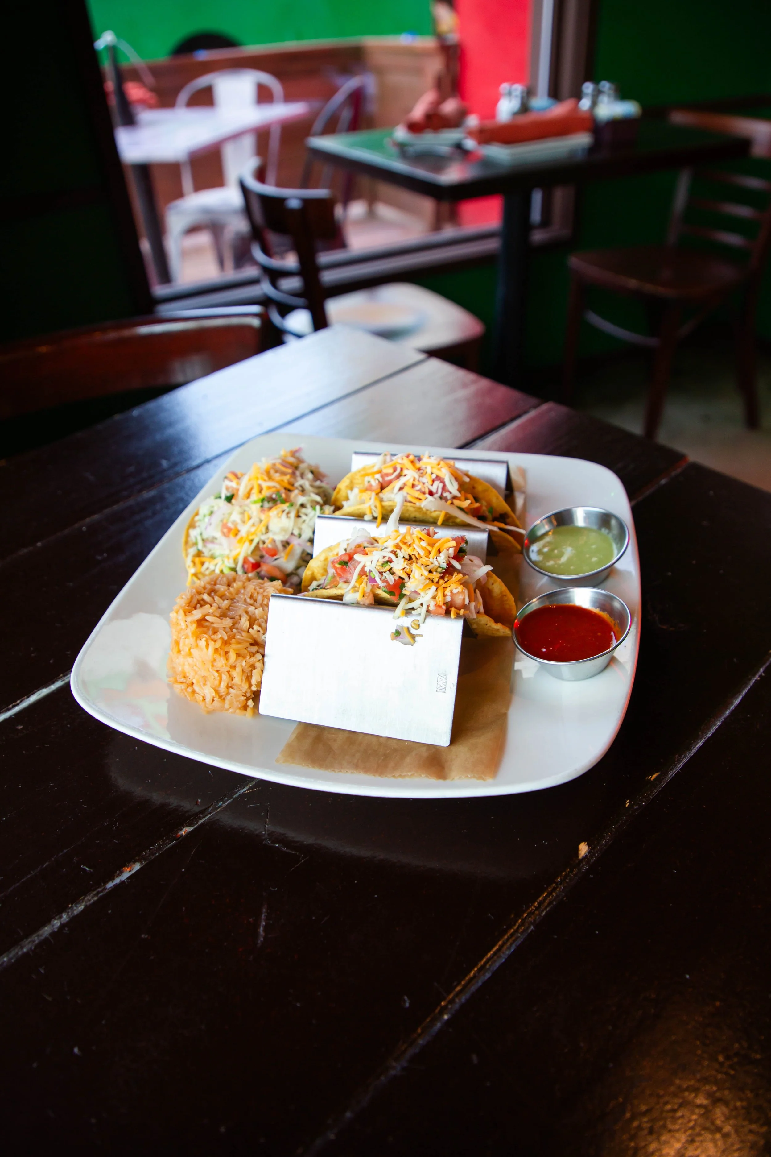 Plate of three tacos topped with shredded cheese, side of rice, and two small containers of green and red sauces on a dark table, restaurant interior in the background.