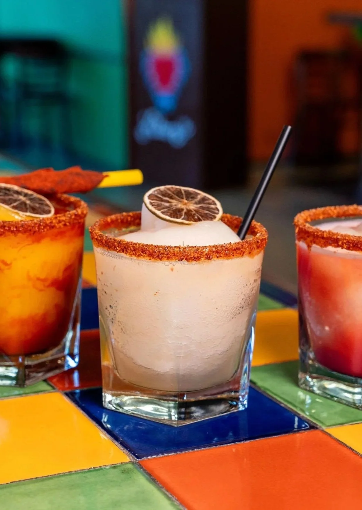 Three colorful cocktails on a table with orange, yellow, green, and blue tiles, garnished with dried lemon slices and chili powder rims.