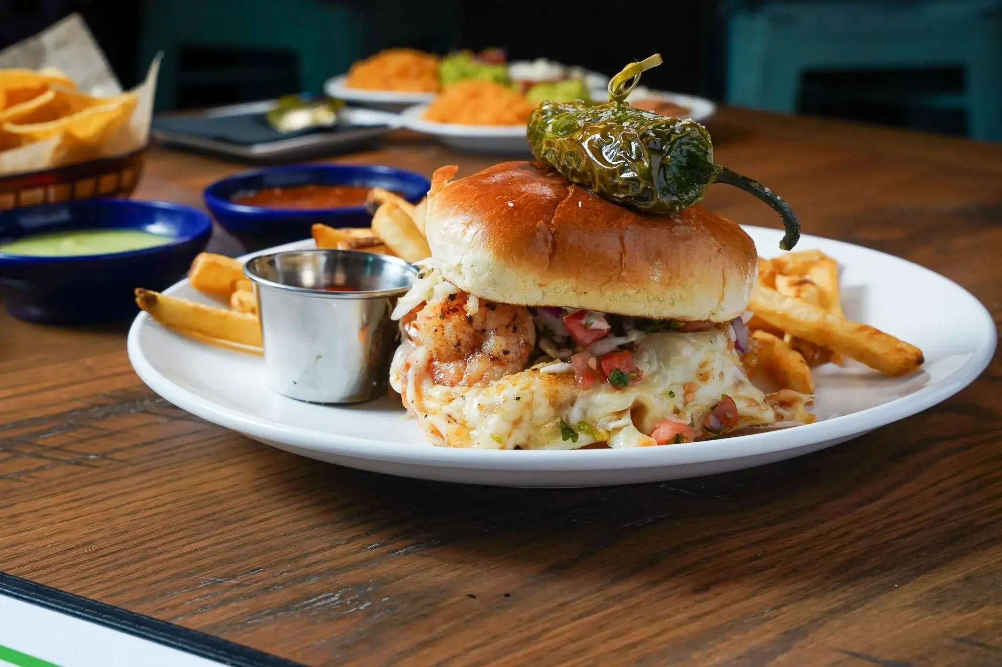 Close-up of a burger with a jalapeño pepper on top, served with fries and various dipping sauces on a white plate.