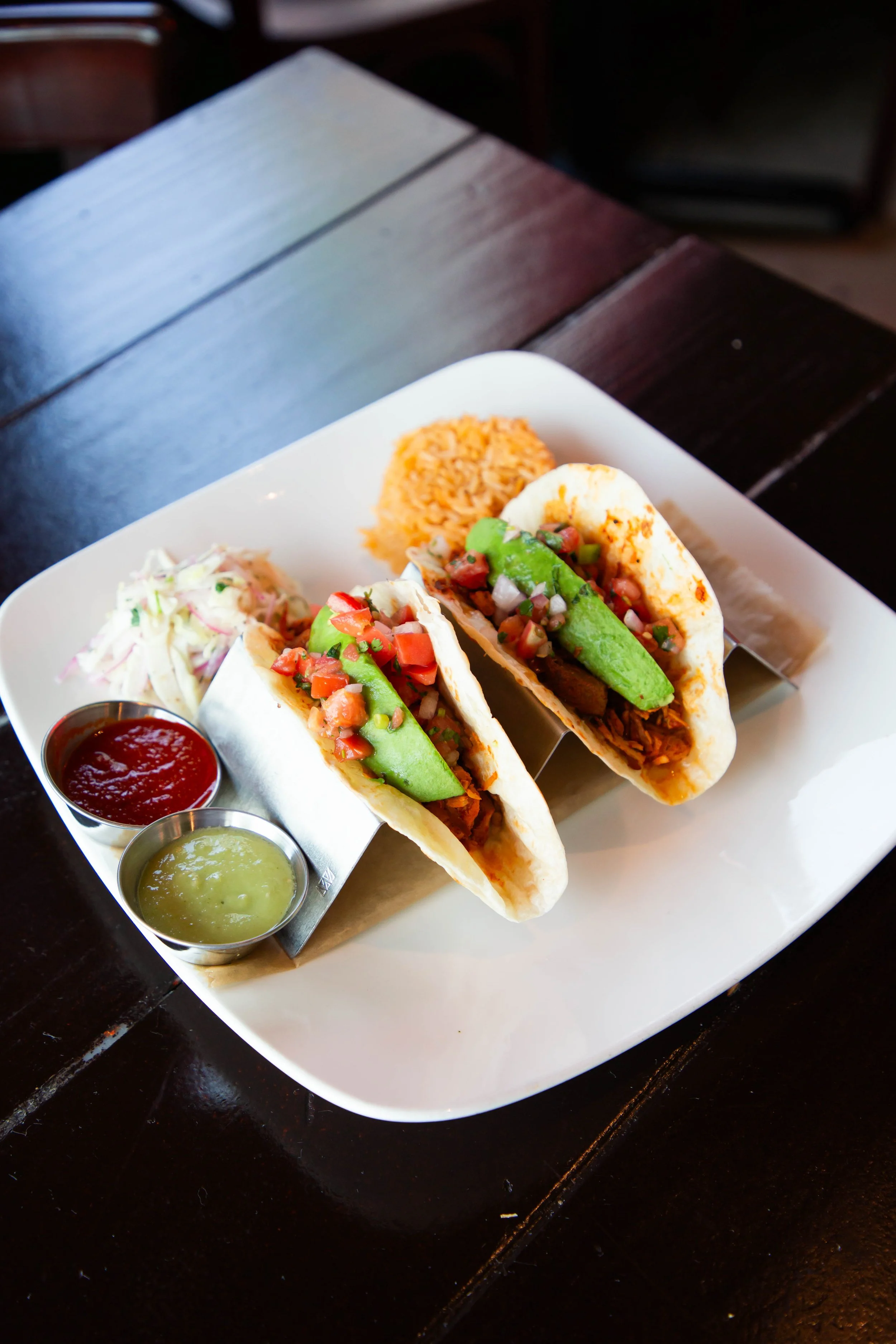 Close-up of two soft tacos filled with shredded meat, chopped tomatoes, onions, and chopped green chilies, topped with salsa and accompanied by sides of coleslaw and rice on a white plate, on a dark wooden table.