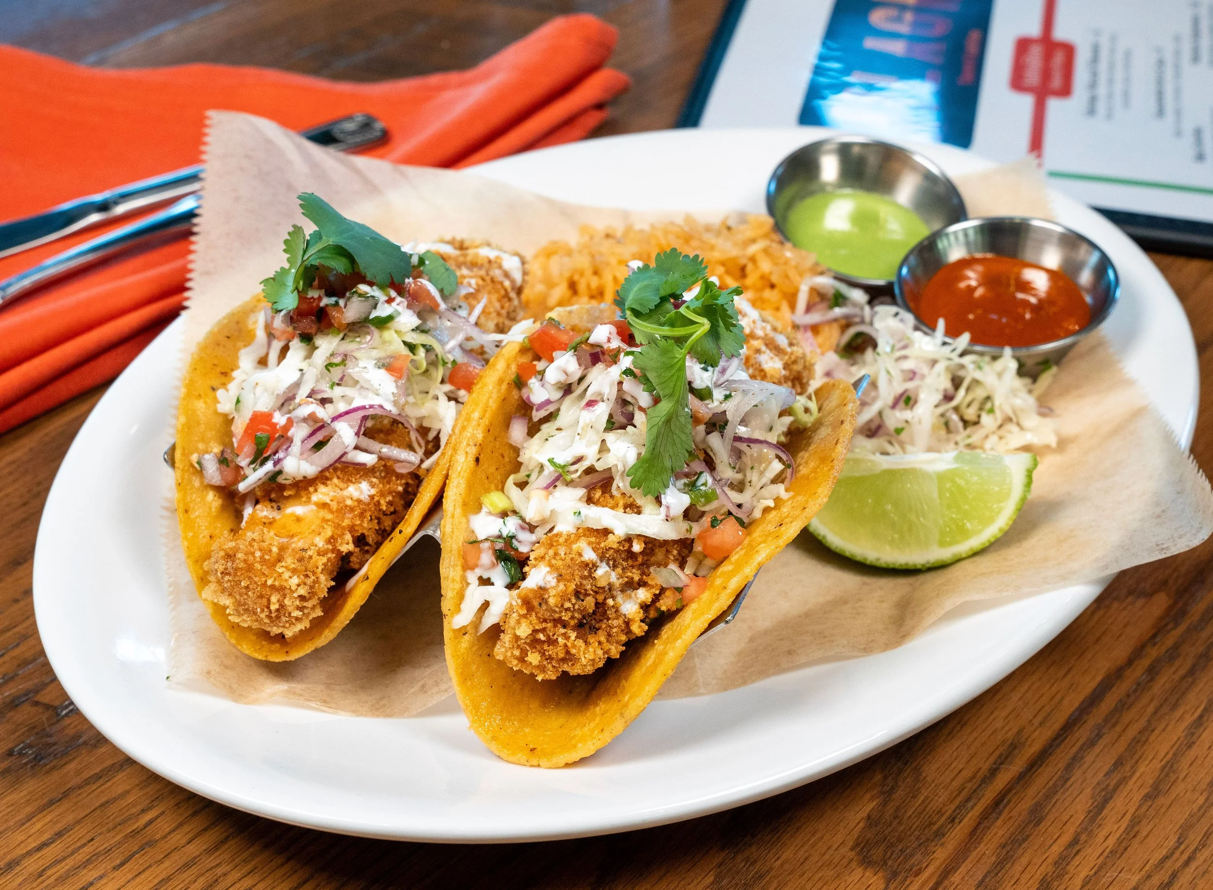 Three crispy fish tacos with shredded lettuce, diced tomatoes, and cilantro served on a white plate with lemon wedges, accompanied by green and red salsa in small metal bowls.