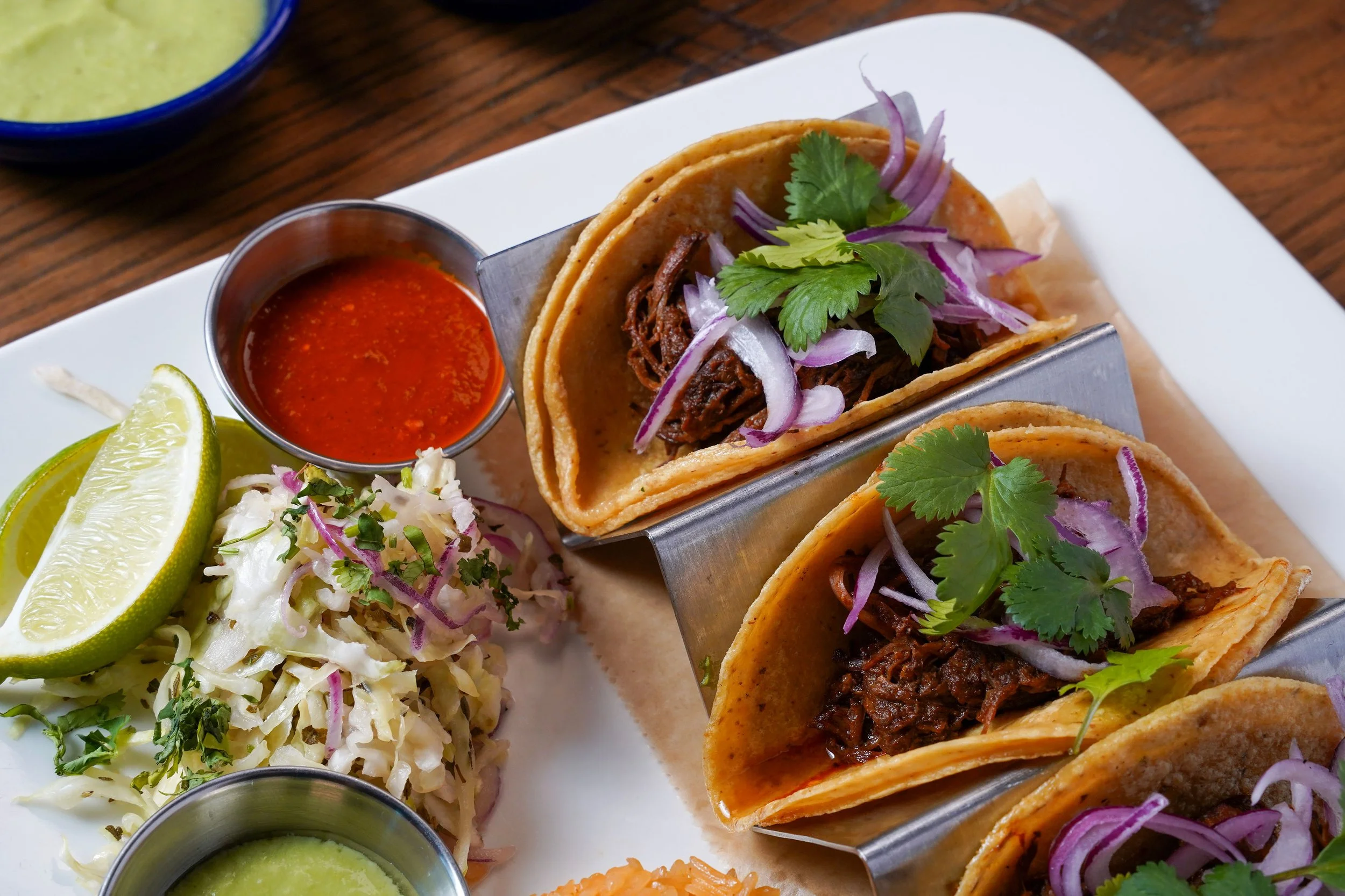 Close-up of three beef tacos topped with cilantro and sliced onions on a white plate, accompanied by lime wedges, shredded cabbage salad, and bowls of green salsa and red salsa.