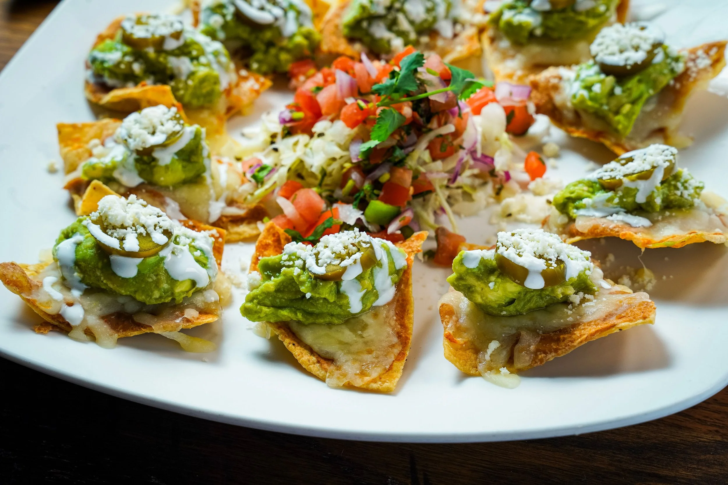 Twelve crispy tortilla nachos topped with guacamole, sour cream, shredded cheese, and sliced jalapenos arranged around a central pile of chopped tomatoes, red onions, and cilantro on a white plate.