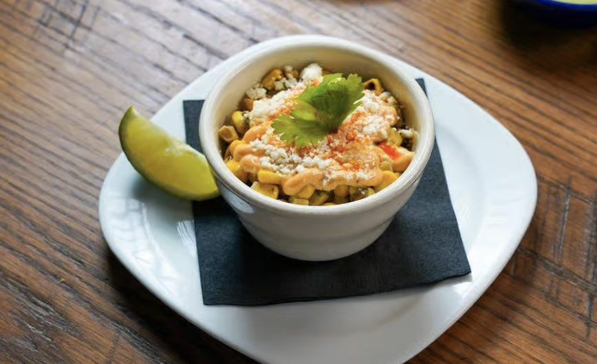 A bowl of Mexican street corn salad garnished with cheese, cilantro, and shredded cheese, served with a lime wedge on a white plate on a wooden table.
