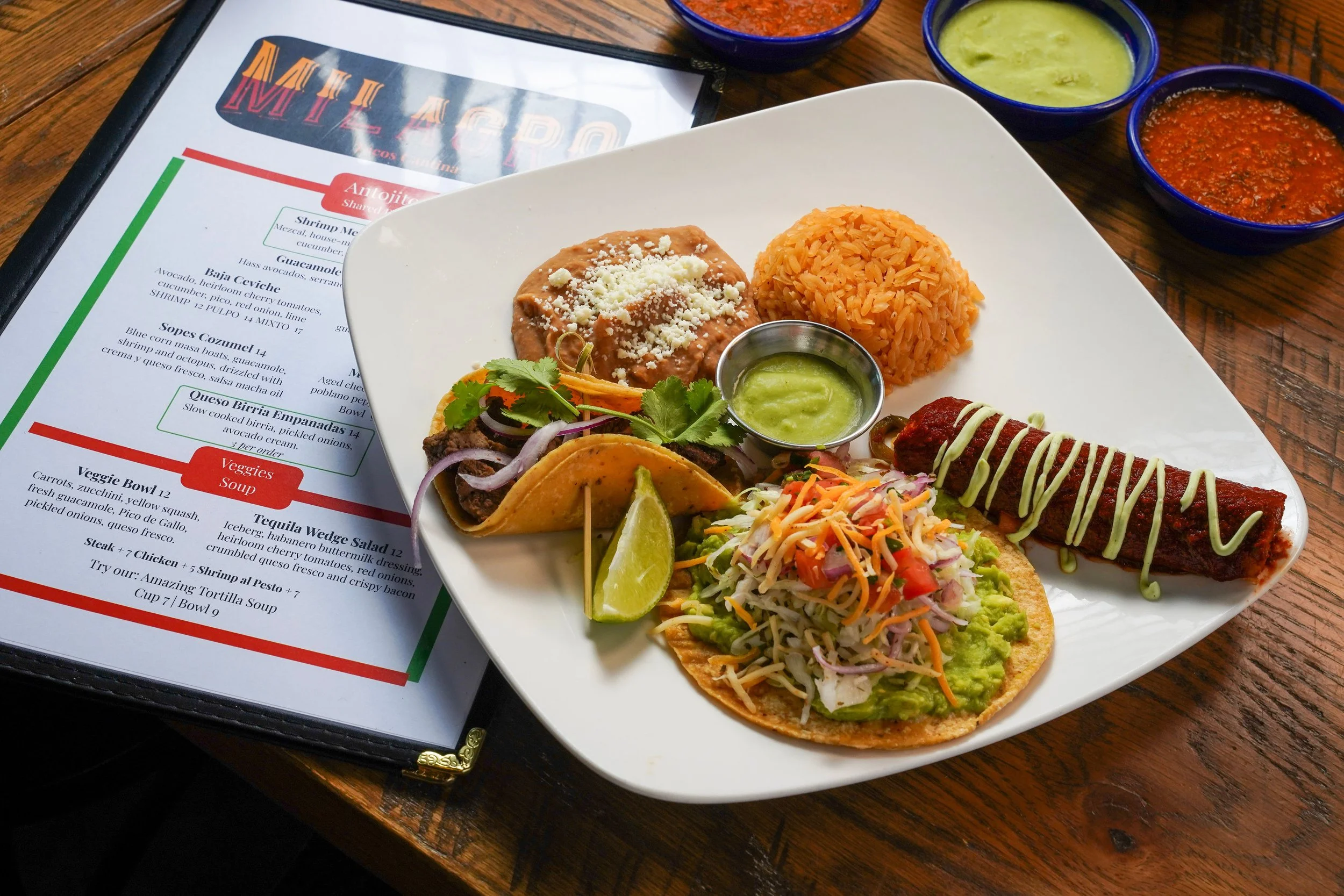 A plate of Mexican food including a taco with guacamole, shredded vegetables, and lime, alongside an enchilada topped with cheese, a serving of Mexican rice, and small bowls of green and red salsa on a wooden table, with a menu visible in the background.