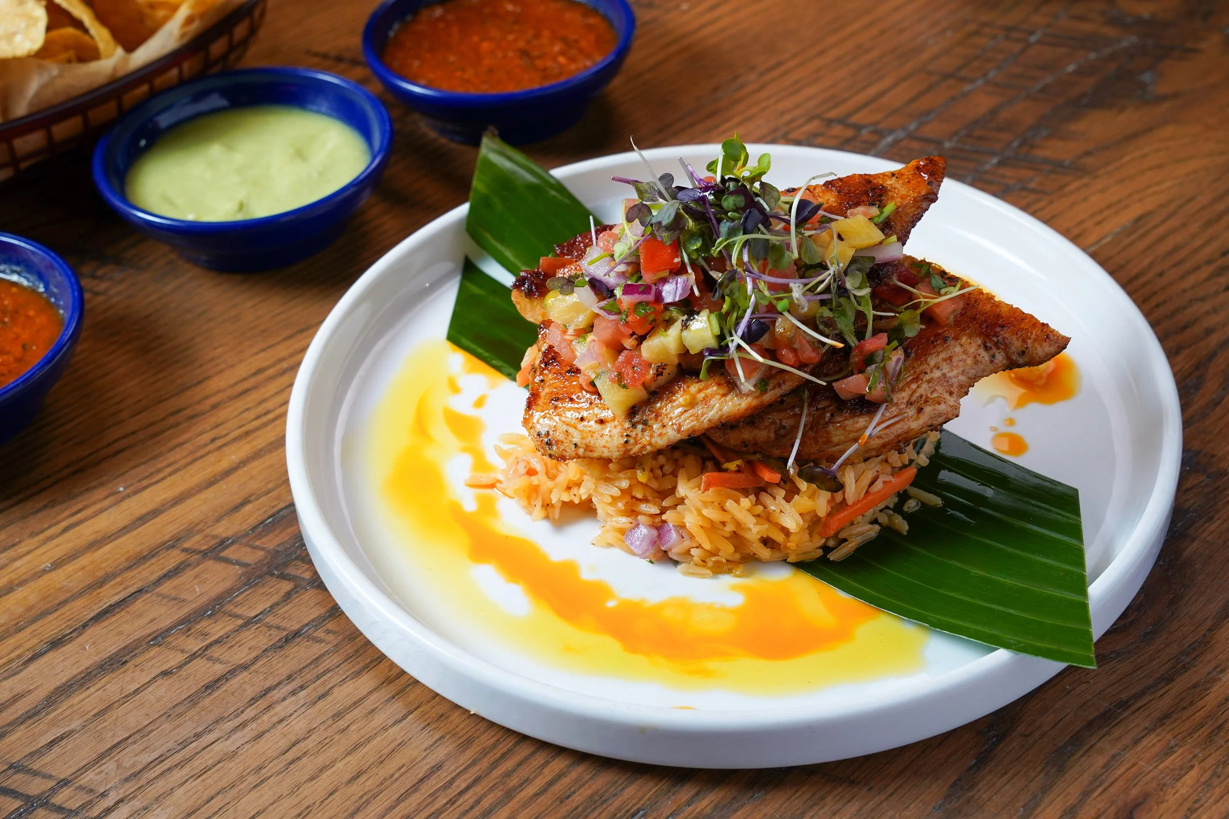 A white plate with grilled fish topped with a colorful vegetable salsa, served over seasoned rice, with microgreens and a green leaf garnish, on a wooden table. Three small bowls of sauces and a basket of chips are in the background.