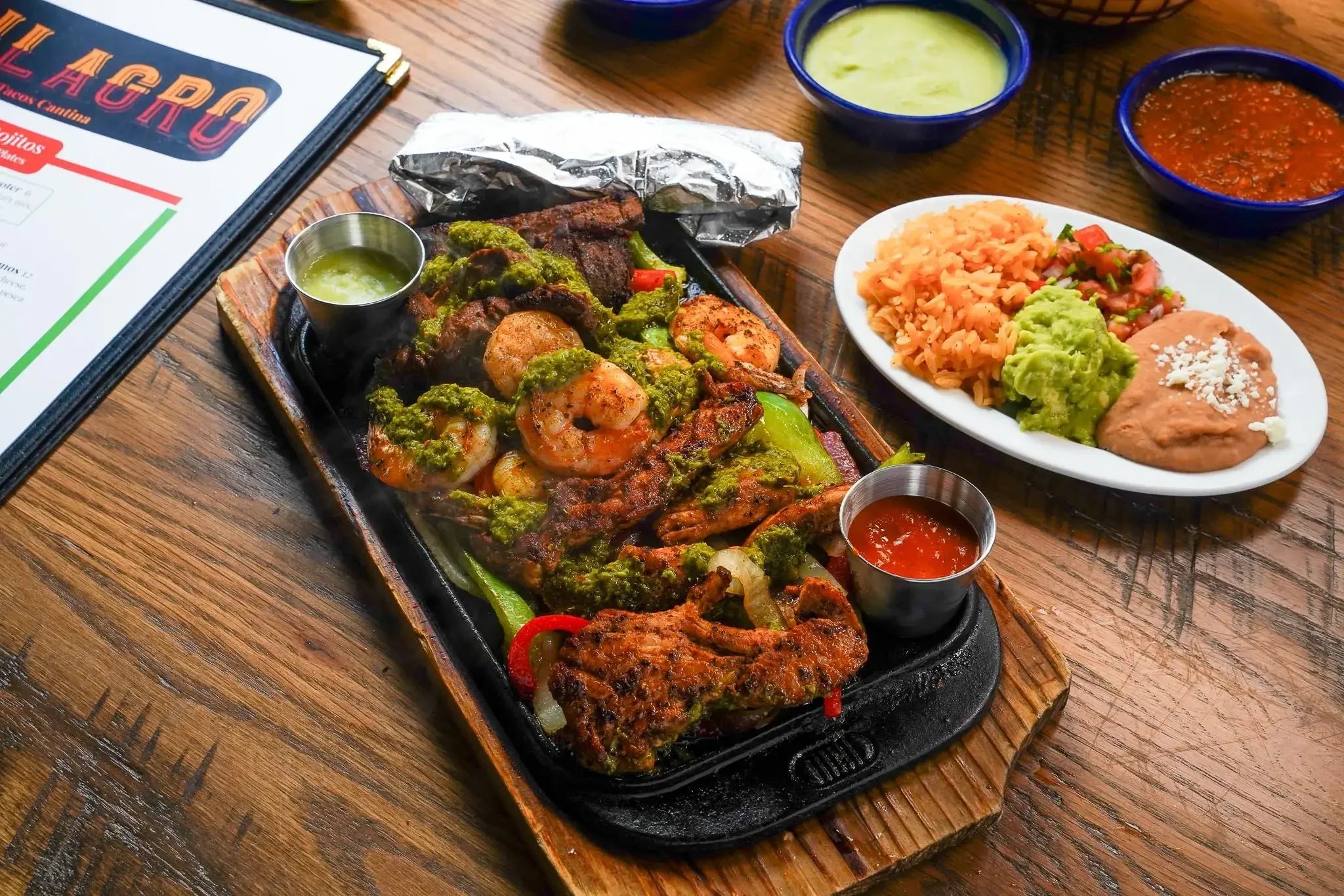 Plate of grilled shrimp with green sauce, served with small cups of additional sauces, on a sizzling skillet. A side plate of Mexican rice, guacamole, pico de gallo, and a small portion of refried beans topped with cheese. Bowls of green and red salsa in the background.