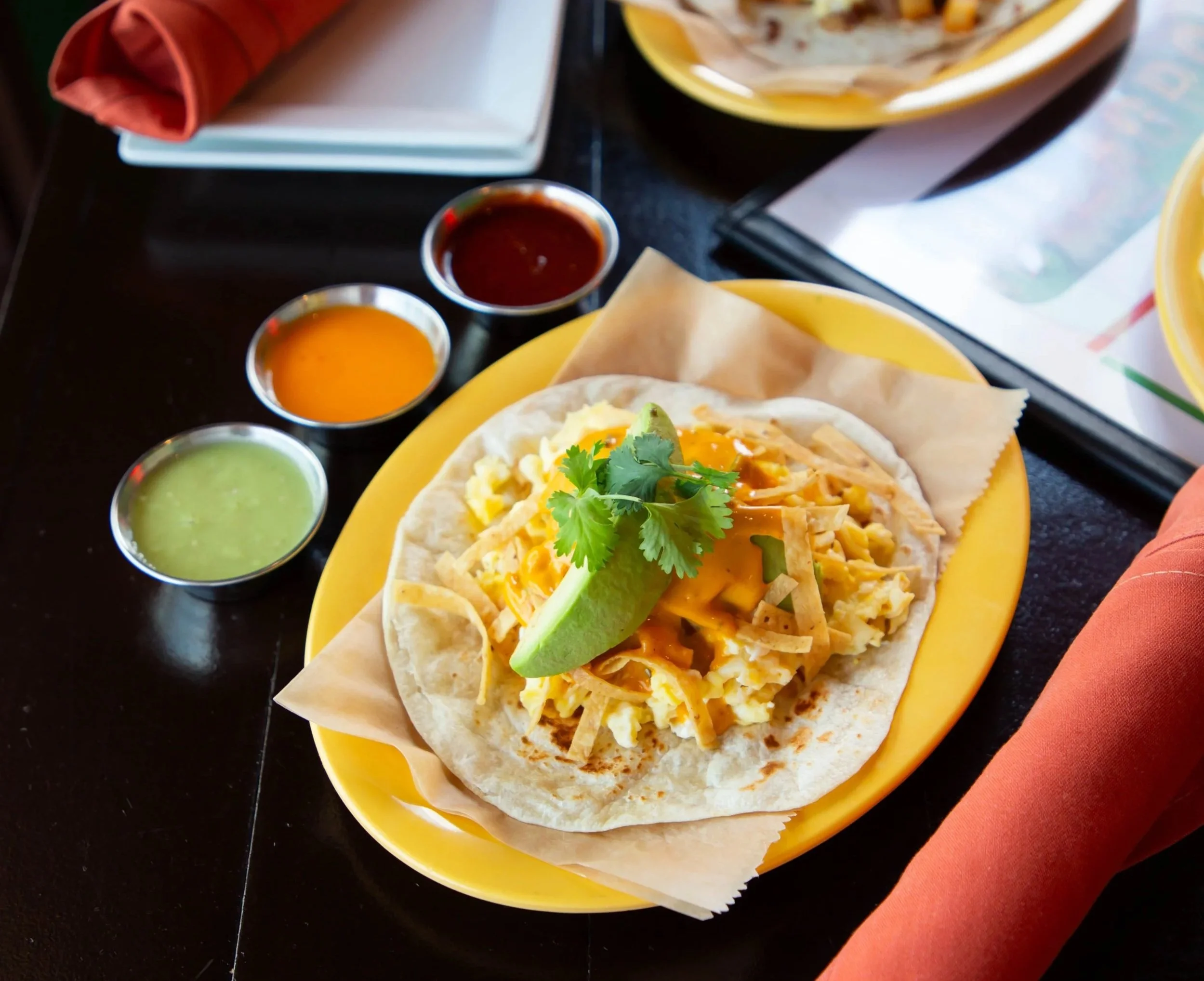 Close-up of a breakfast taco filled with scrambled eggs, shredded cheese, and cilantro, served on a yellow plate with three small containers of salsa and guacamole, with a black table and some napkins in the background.