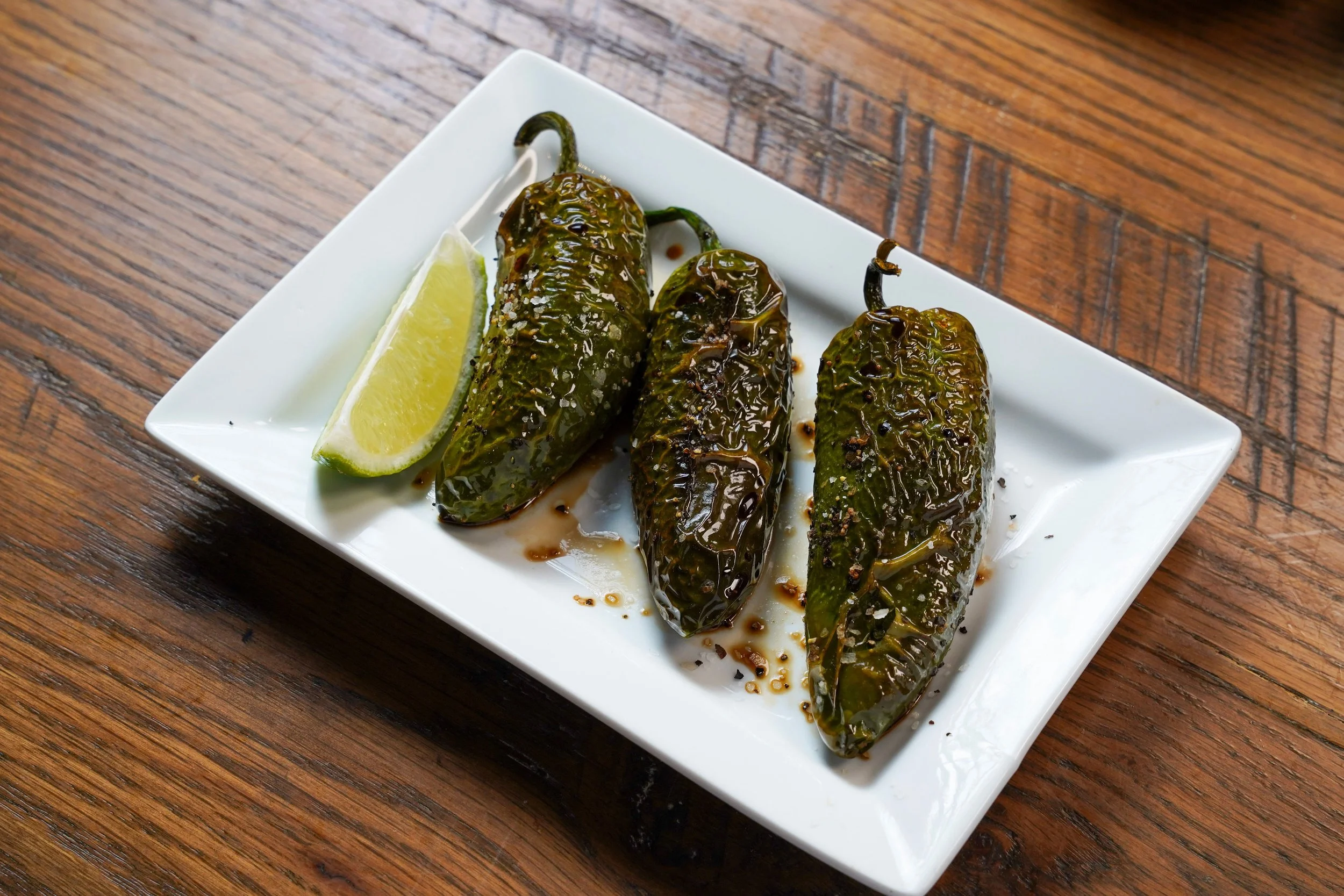 Three roasted hatch green chiles on a white square plate with a lemon wedge on the side, placed on a wooden table.
