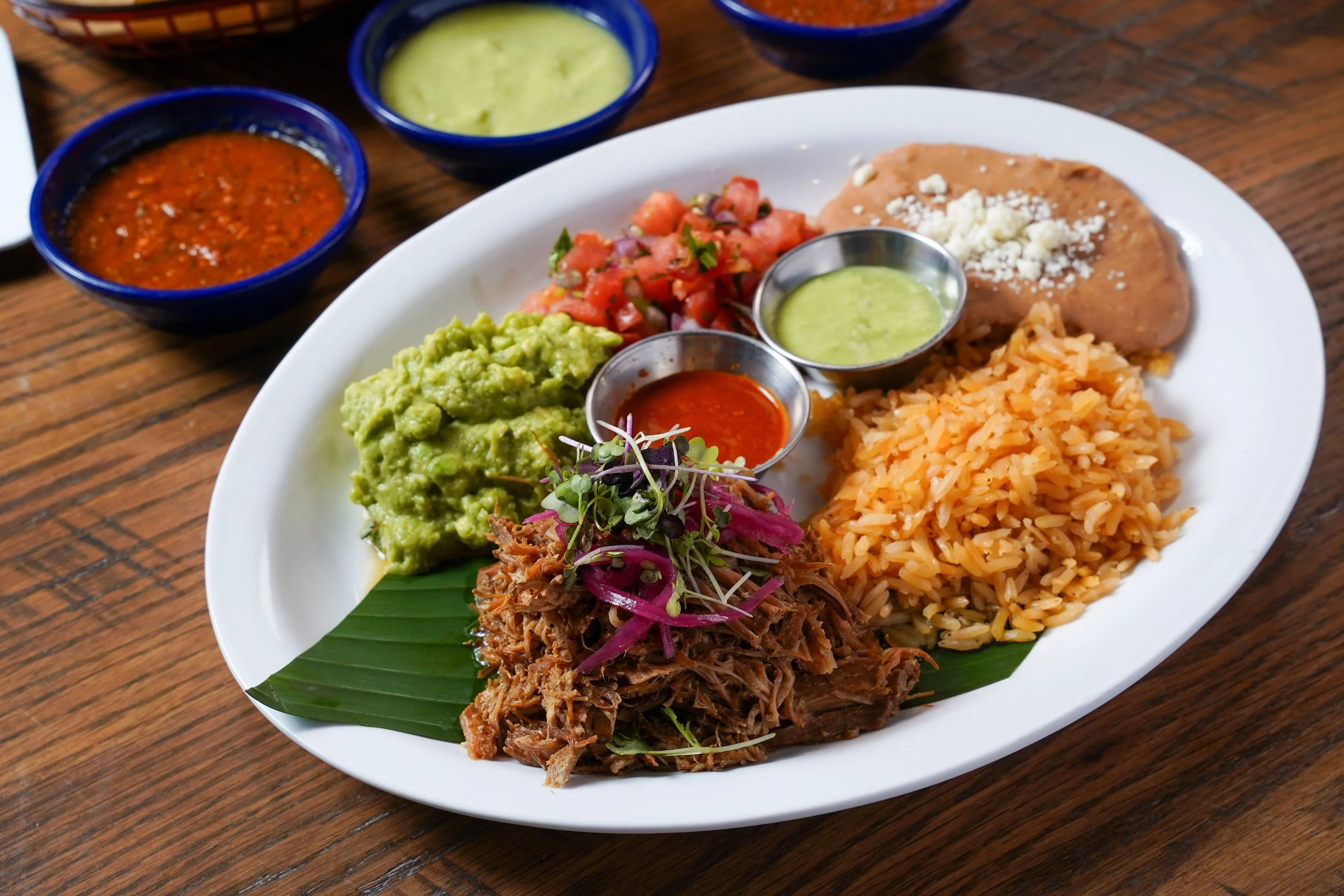 Plate of Mexican food with rice, shredded beef, guacamole, pico de gallo, and various sauces, accompanied by bowls of salsa and guacamole on a wooden table.