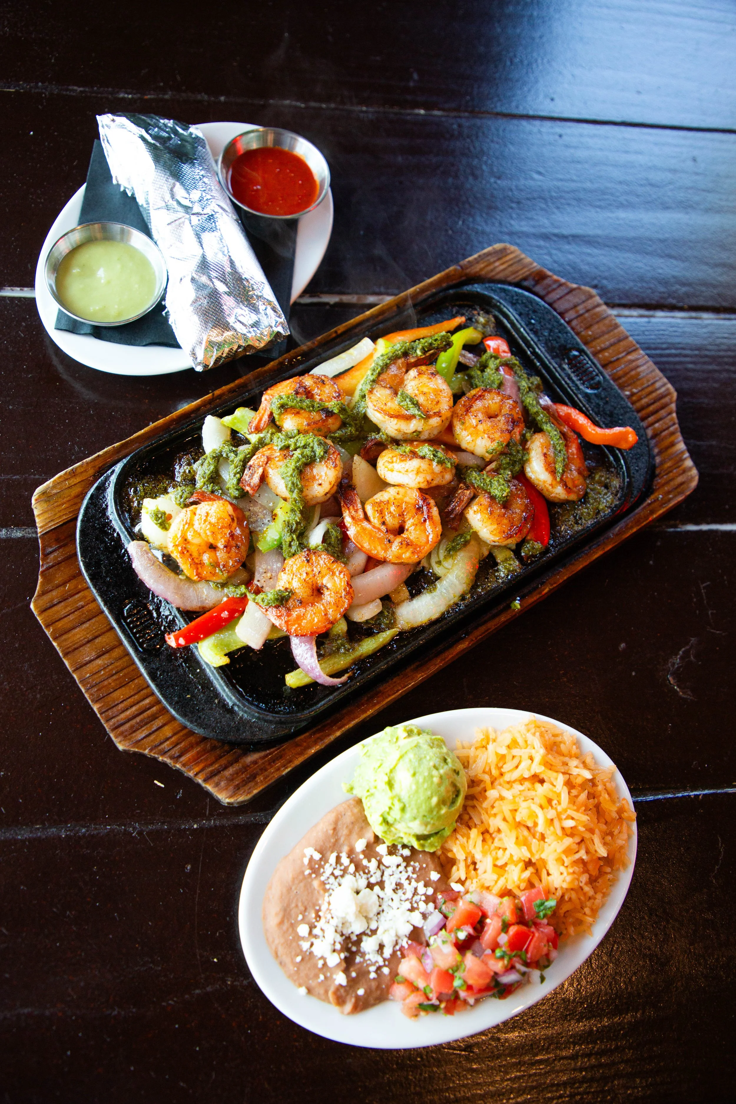 A serving of shrimp fajitas on a sizzling hot plate with vegetables, accompanied by two small bowls of salsa and guacamole, and a side of rice and refried beans topped with cheese, served on a dark wooden table.
