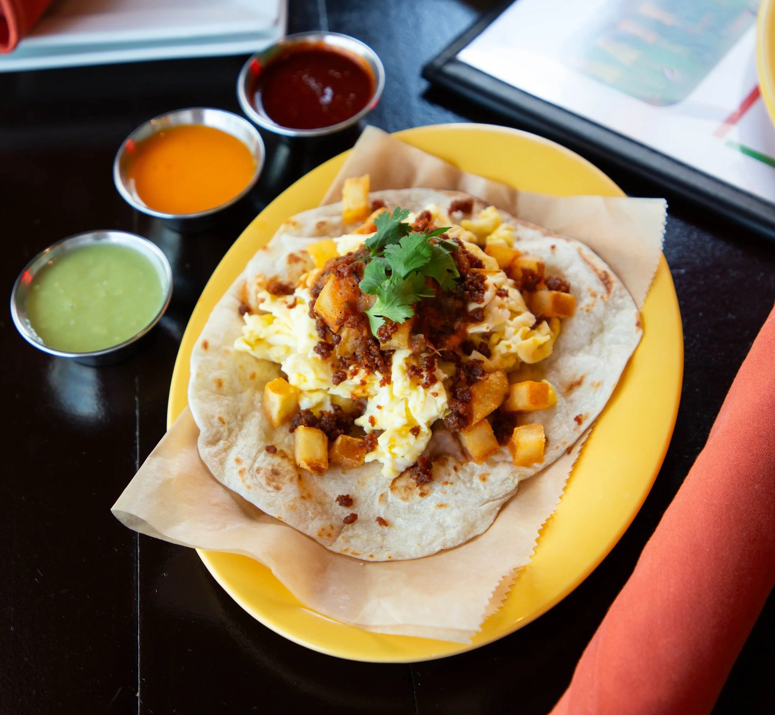 Close-up of a taco on a yellow plate with three small bowls of sauces beside it, one green, one red, and one orange, on a black table.