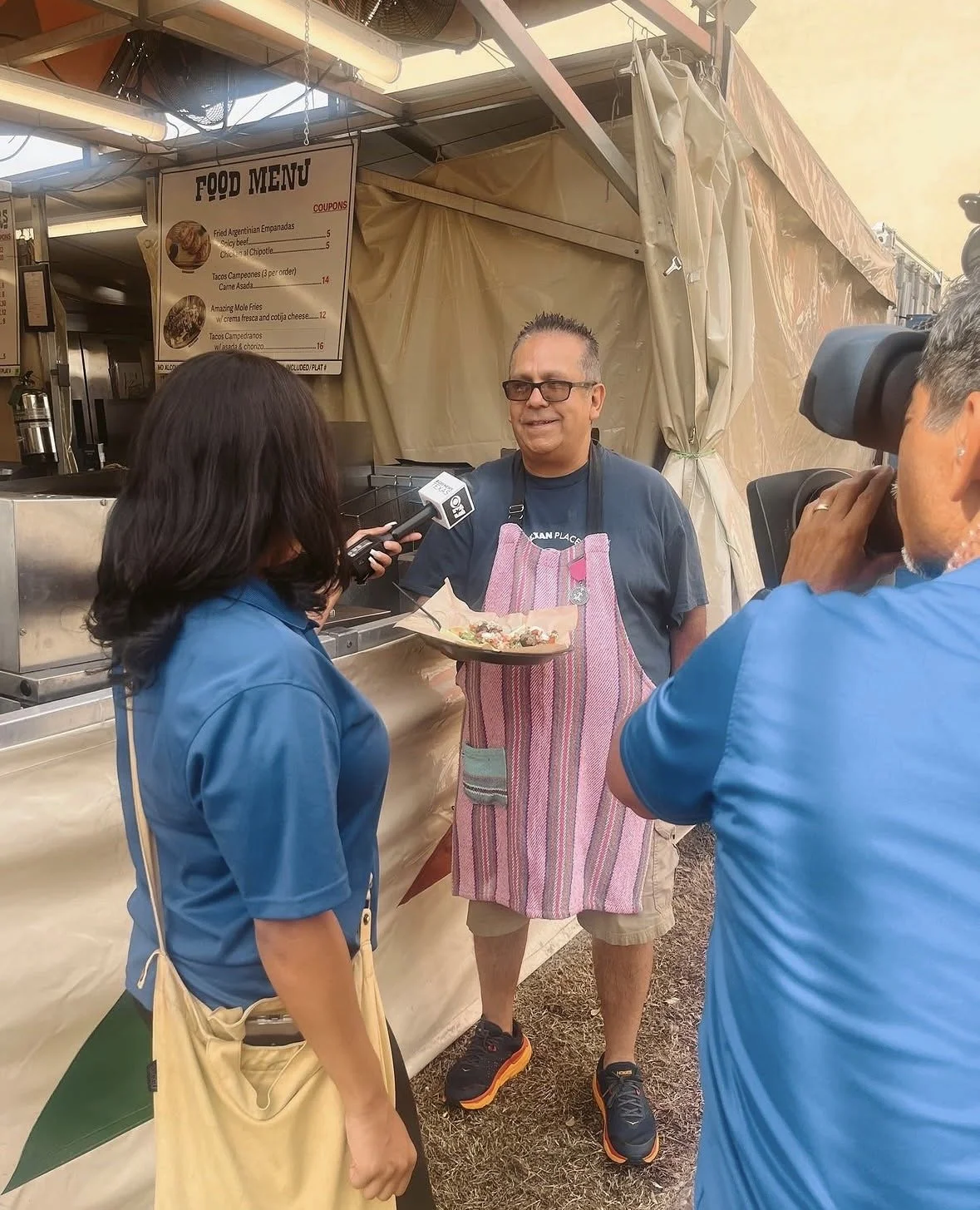 A man wearing a pink and white striped apron is being interviewed at a food stand. A woman with dark hair, wearing a blue shirt, is holding a microphone, and a cameraman in a blue shirt is filming the scene. The food stand has a menu board in the background with various items listed.