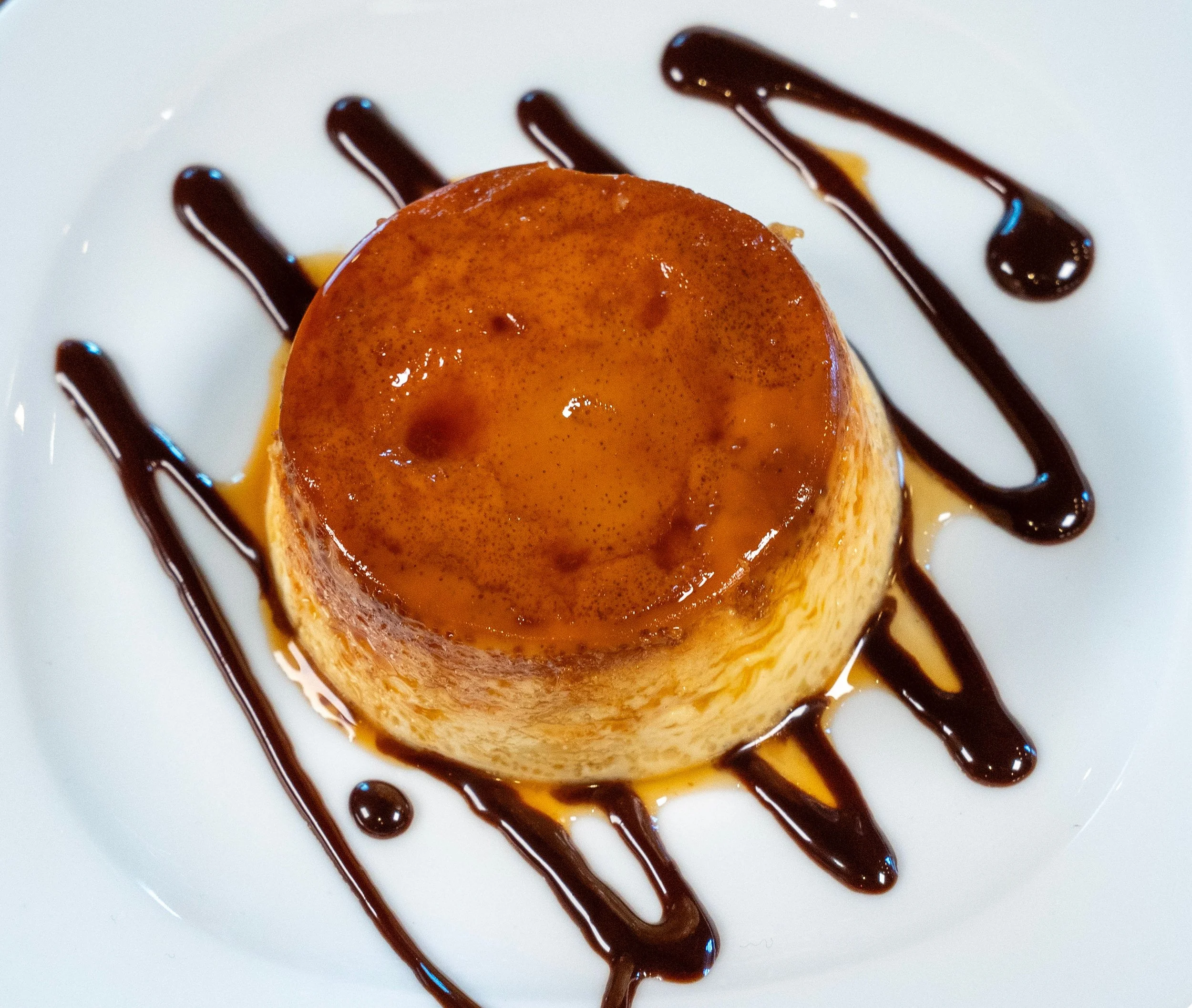 A dessert featuring a small flan with caramel topping, garnished with chocolate and caramel drizzle on a white plate.