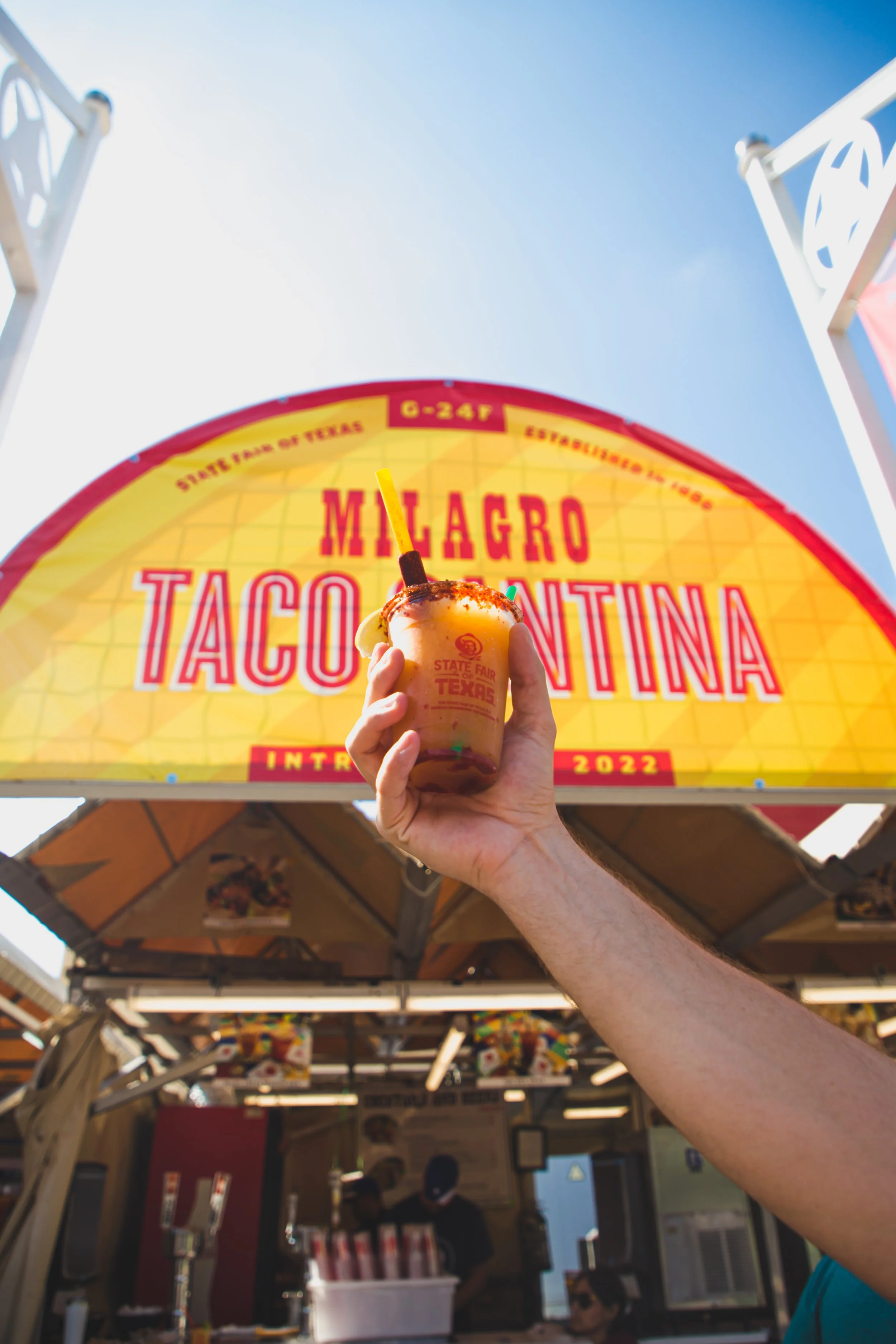 Person holding a drink in front of a yellow sign with red and purple lettering reading 'Milagro Taco Cantina' at an outdoor event.