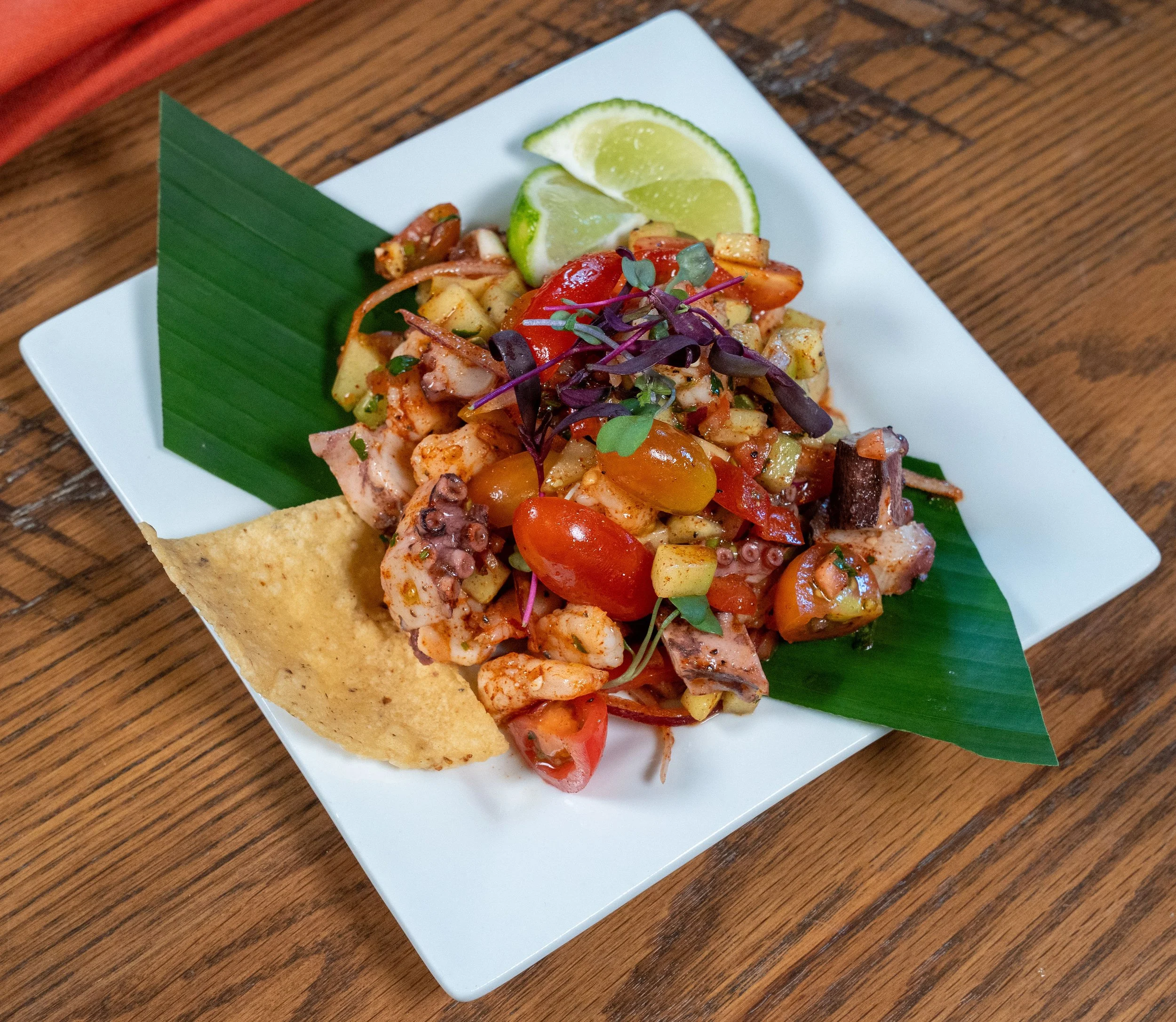 A plate of seafood salad with lime wedges, garnished with microgreens and a banana leaf on a white square plate, on a wooden table.