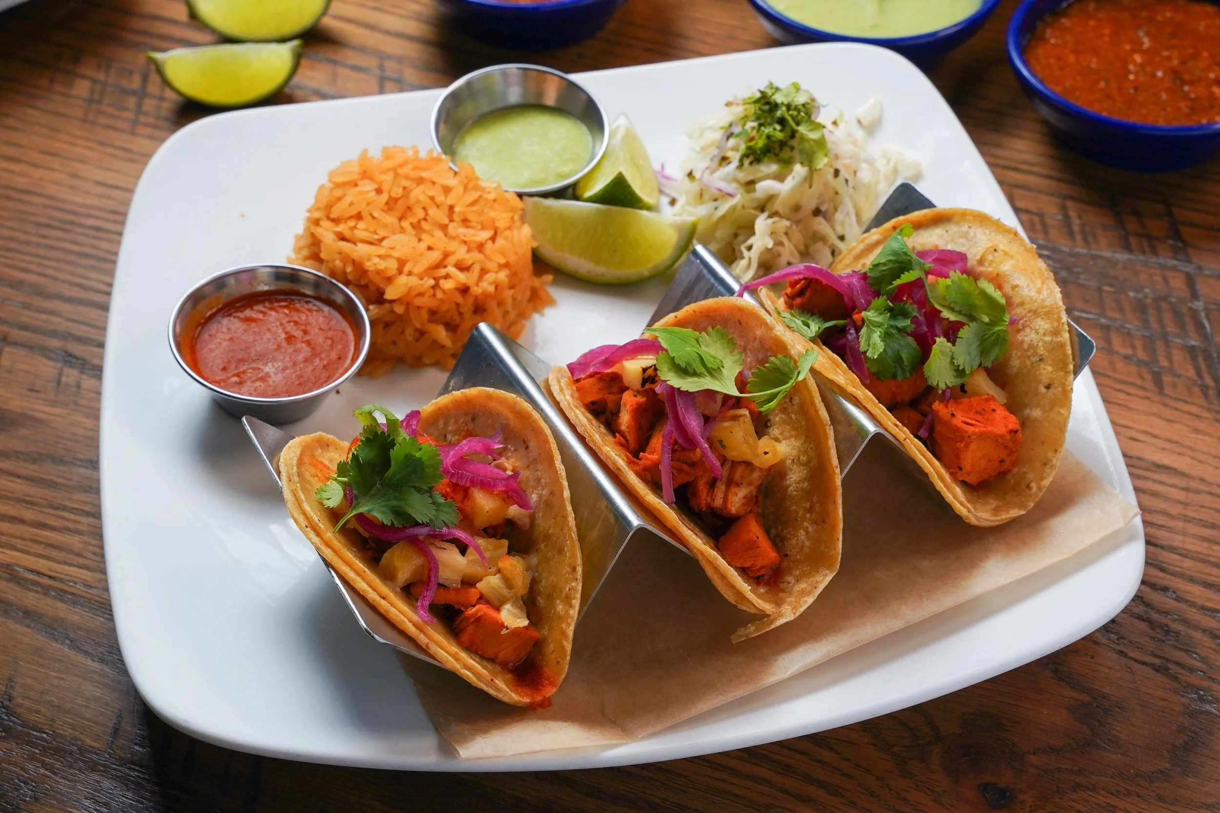 Four tacos filled with seasoned chicken, red onions, and cilantro on a white platter with lime wedges, two small cups of red and green sauces, and a side of rice and cabbage slaw. In the background, there are lime wedges and small bowls of sauces on a wooden table.