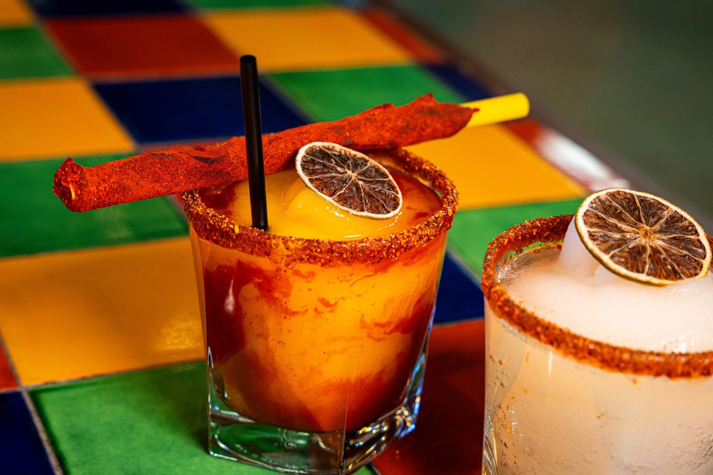 Close-up of two colorful cocktails garnished with dried lemon slices and chili powder rimmed glasses, set against a vibrant, multicolored tiled background.