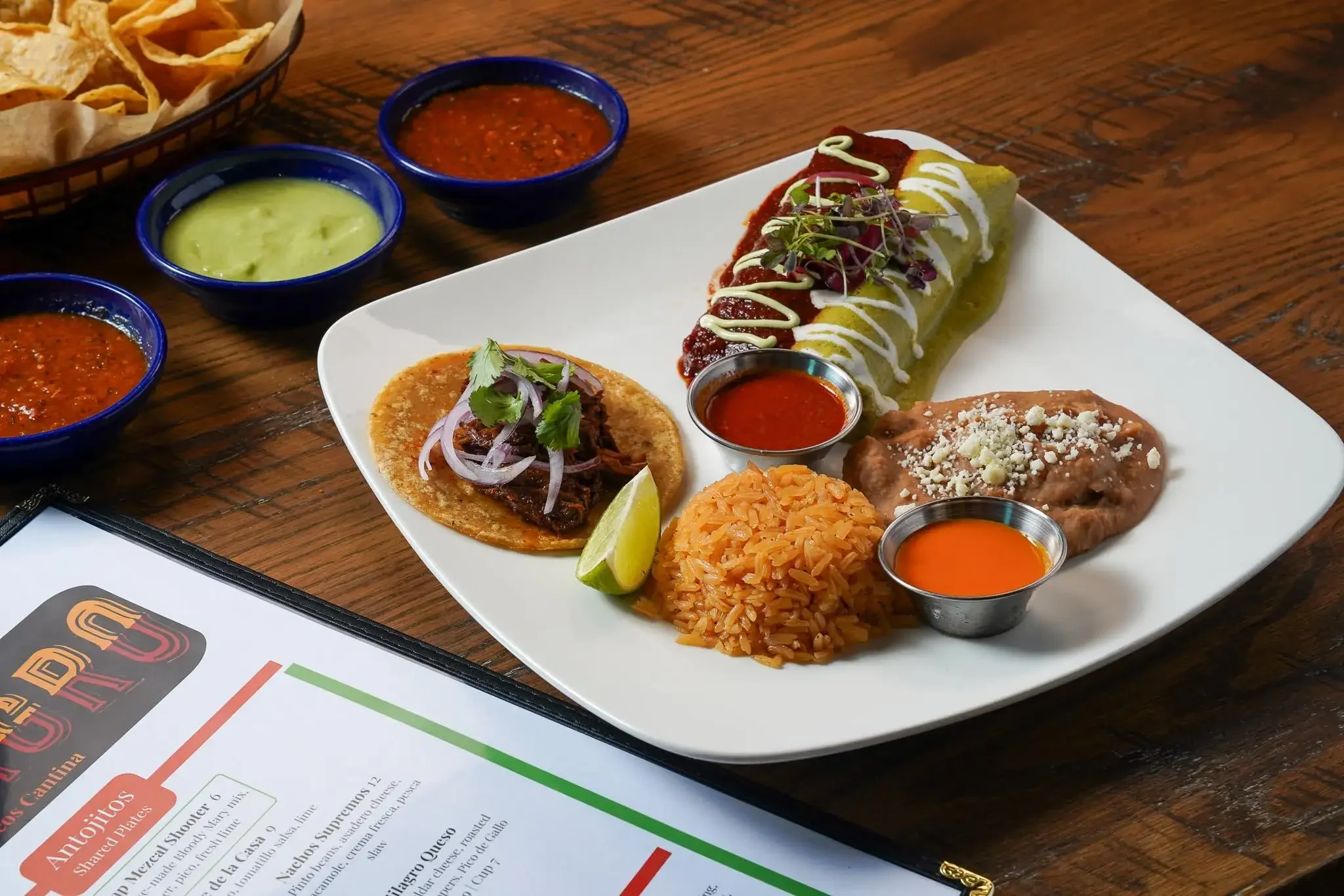 A plate of Mexican food including a stuffed pepper with sauce, Mexican rice, refried beans with cheese, a taco with shredded beef and onions, and a baked tostada with cheese. There are small bowls of red, green, and hot sauce on the side.