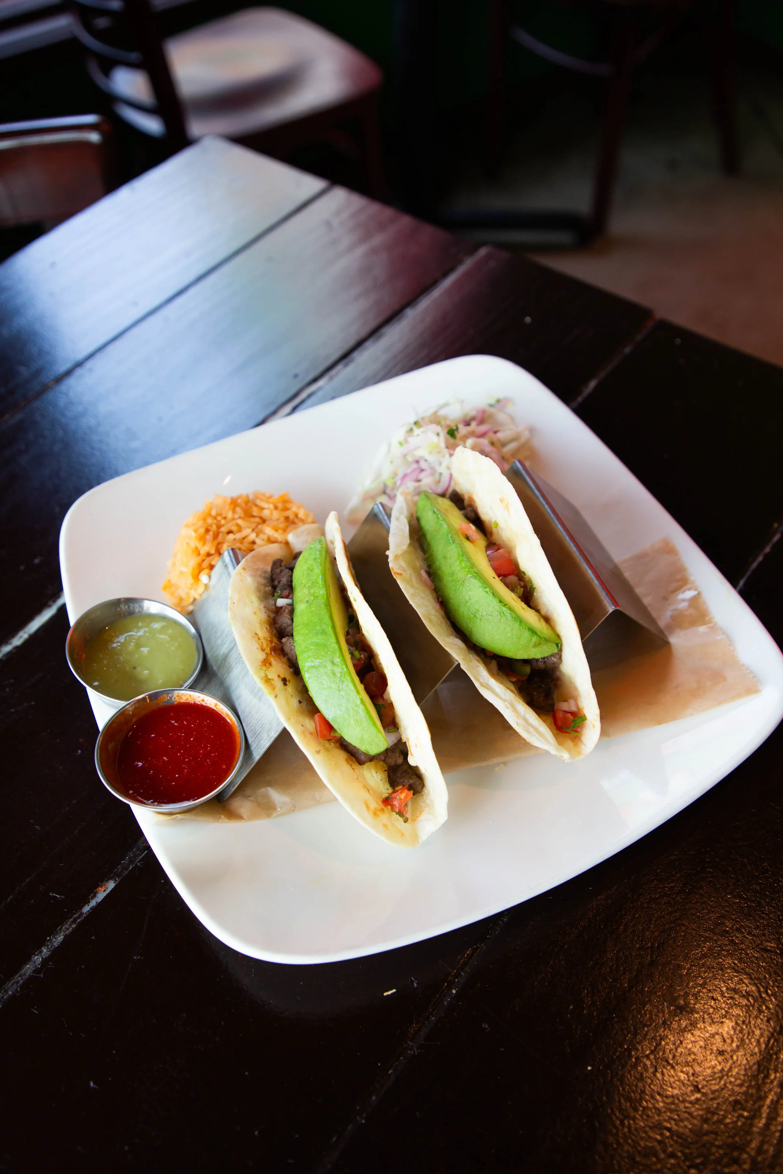 A white rectangular plate with three beef tacos filled with avocado slices, chopped tomatoes, and onions, accompanied by three small cups of salsa and guacamole, on a dark wooden table.