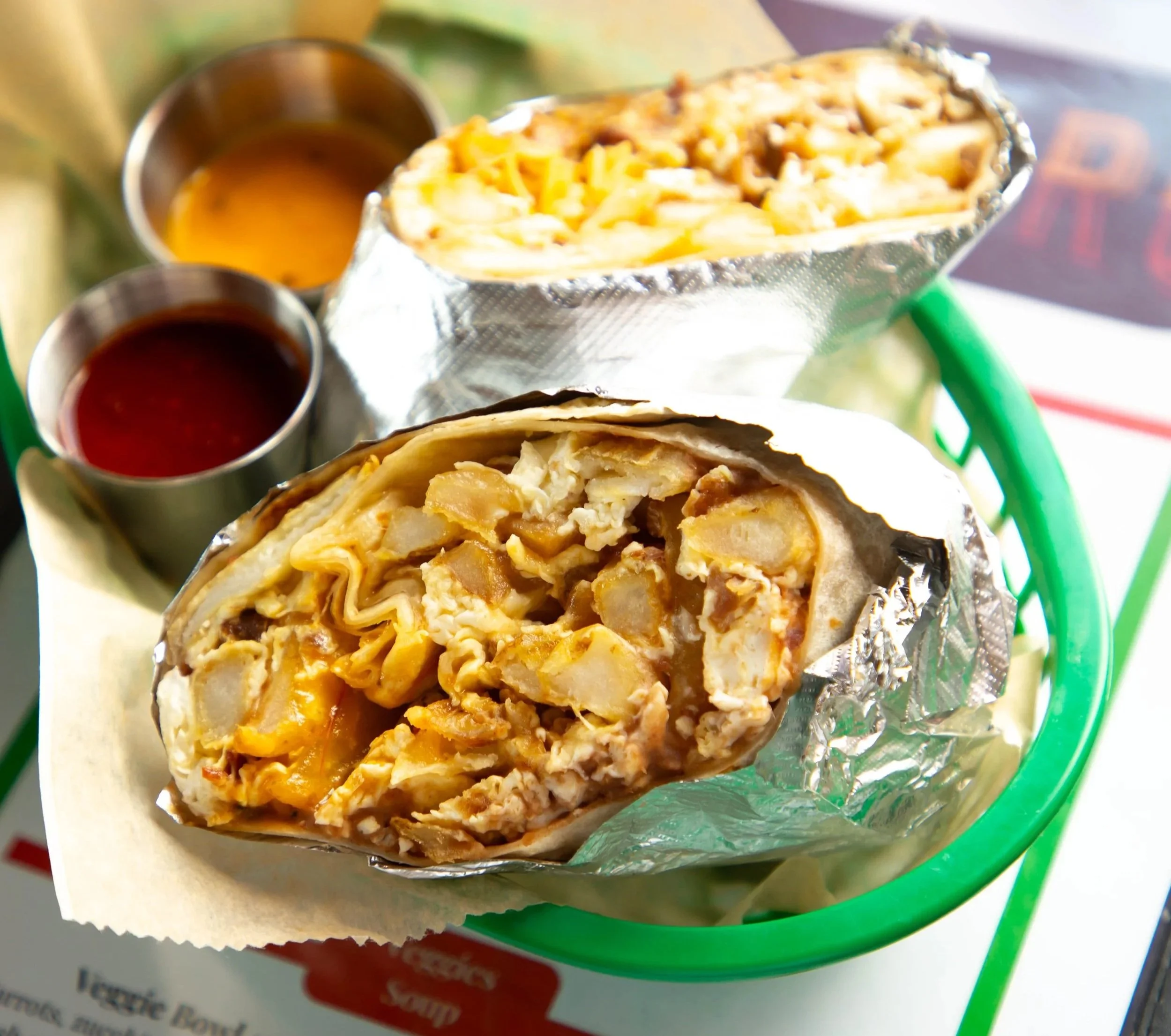 A burrito cut in half showing stuffed ingredients such as meat, cheese, and potatoes, wrapped in foil, with two small cups of sauce nearby on a plate.