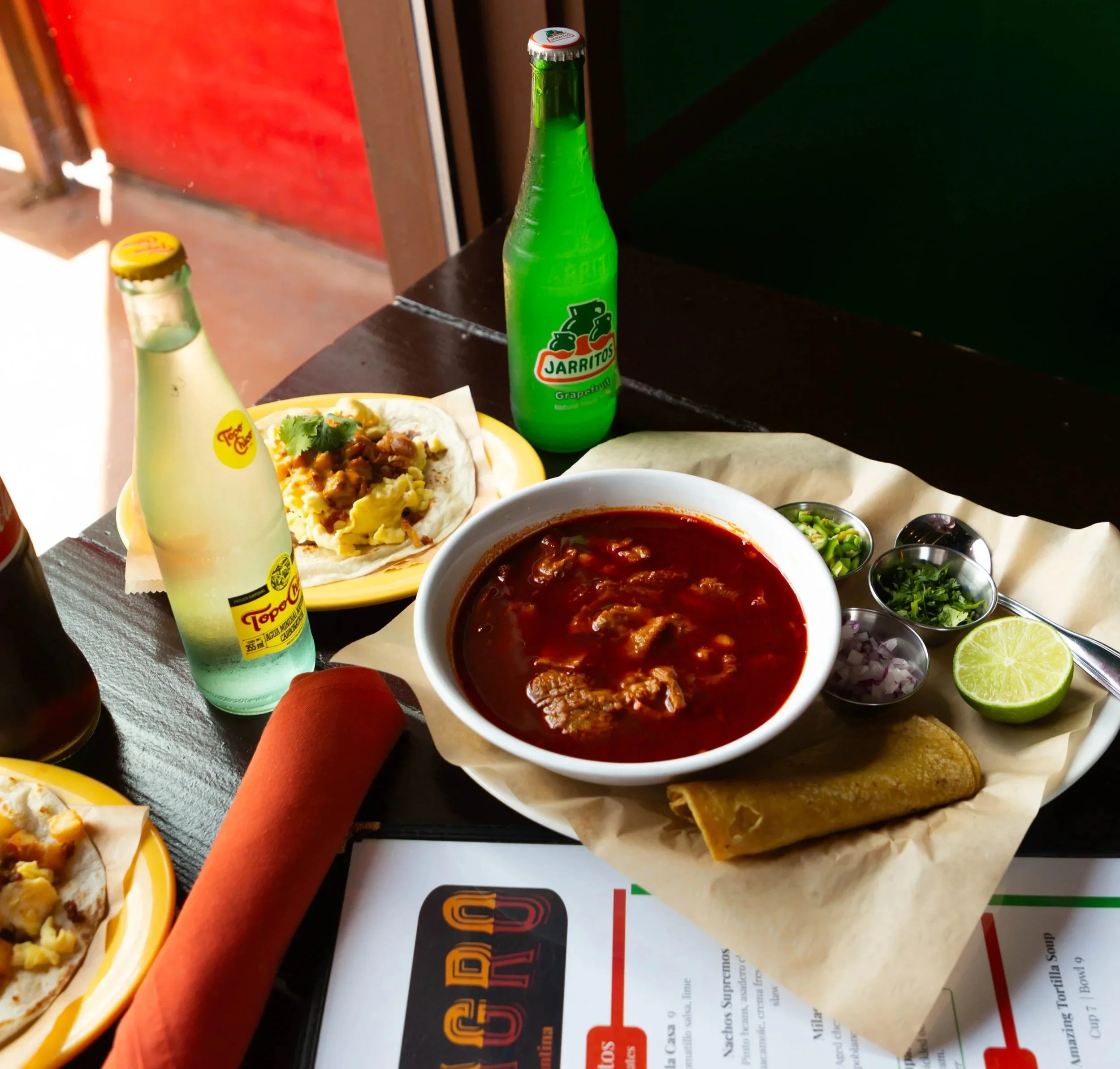 A table with Mexican food including a bowl of red beef chili with vegetables and meat, a yellow plate with tacos filled with meat, cheese, and cilantro, bottles of Jarritos grapefruit soda and Topo Chico mineral water, and small bowls of chopped onions, cilantro, and lime wedges.