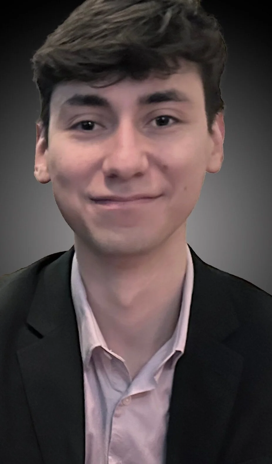 A young man with brown hair wearing a black suit and a light pink shirt, smiling softly at the camera, against a dark gradient background.