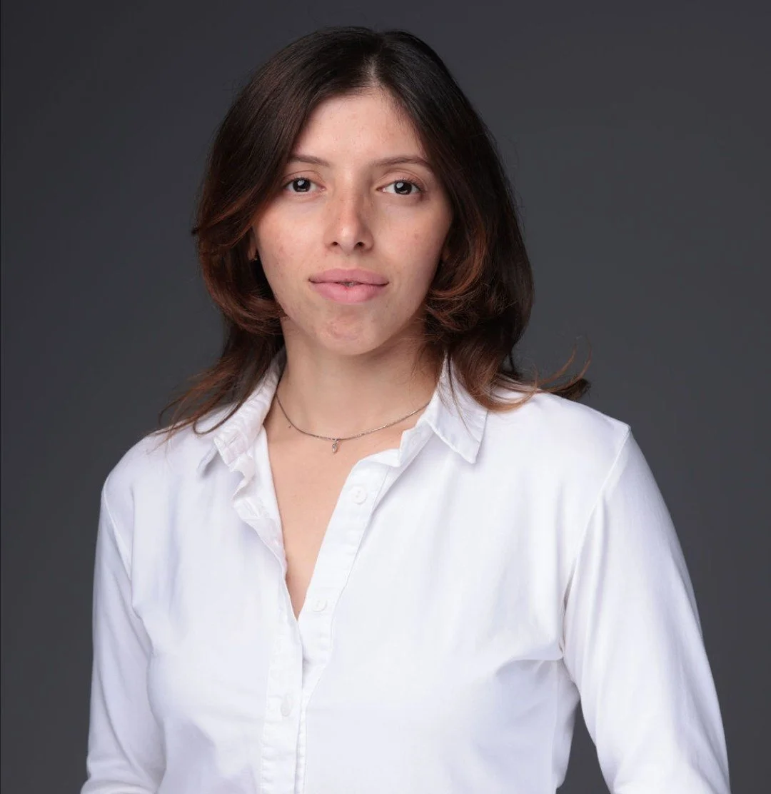 A woman with shoulder-length brown hair wearing a white button-up shirt against a gray background.
