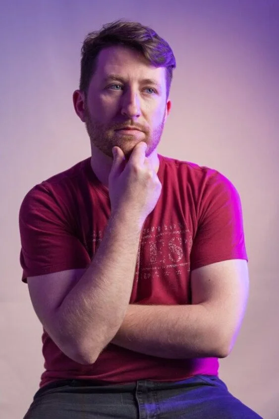 A man with short brown hair and a beard is sitting with his right hand resting on his chin, looking thoughtful. He is wearing a red t-shirt and dark pants, with a purple and pink gradient background.