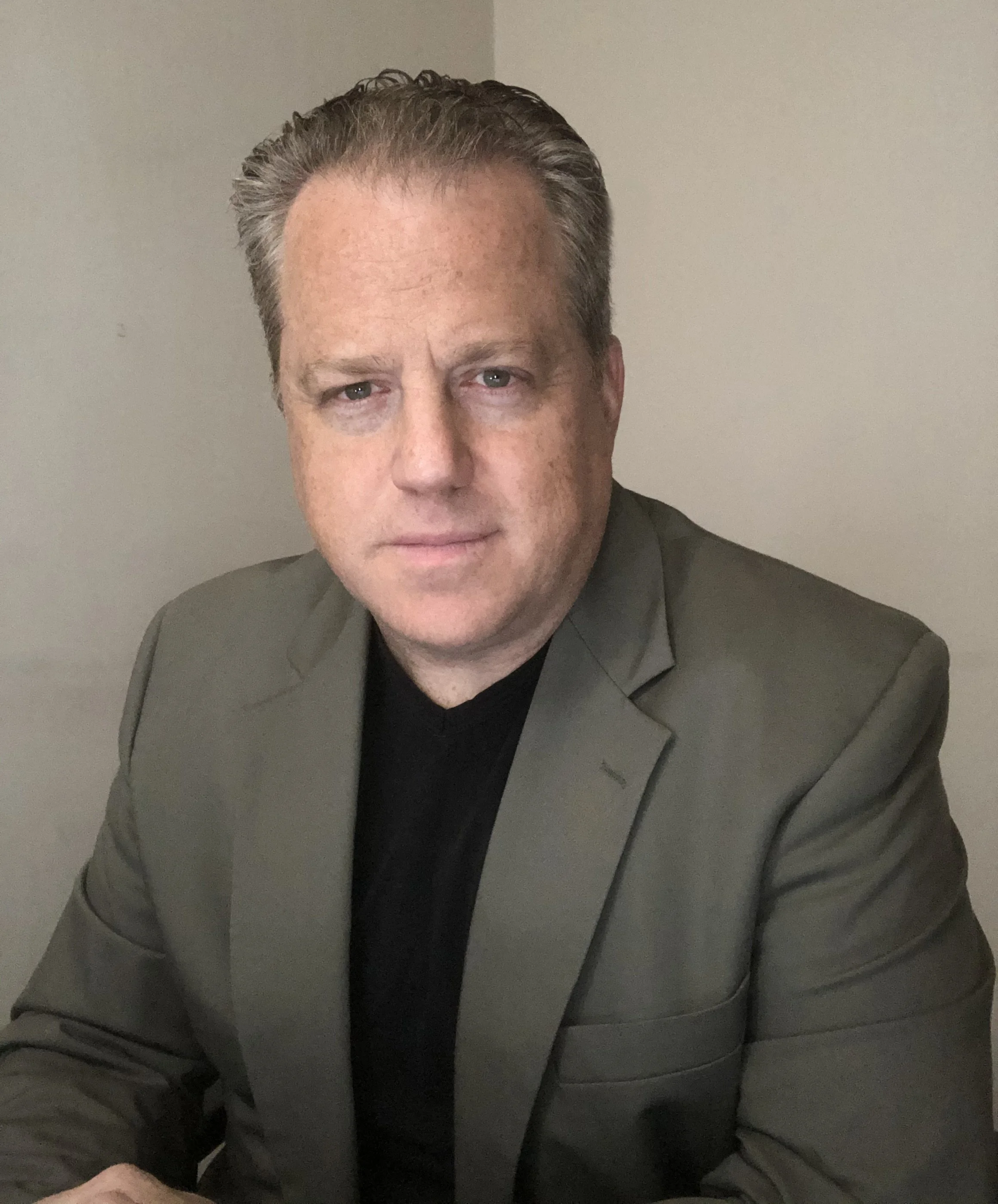Rob Pohle, post production supervisor wearing a gray blazer over a black shirt, posing for a portrait against a plain beige wall.