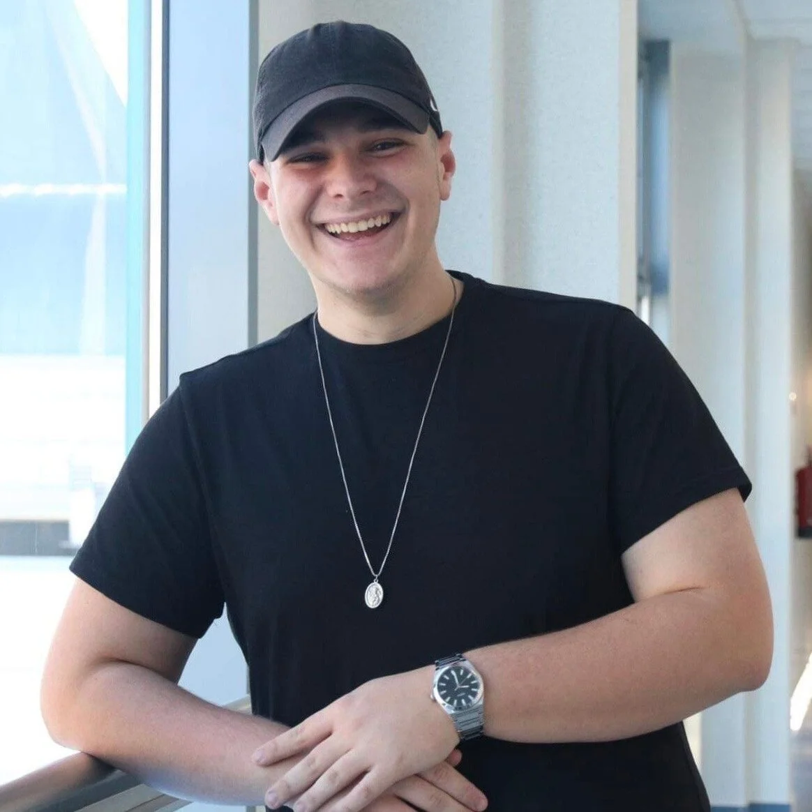 A young man wearing a black t-shirt, a black baseball cap, a silver watch, and a necklace, smiling and leaning against a railing inside a building.