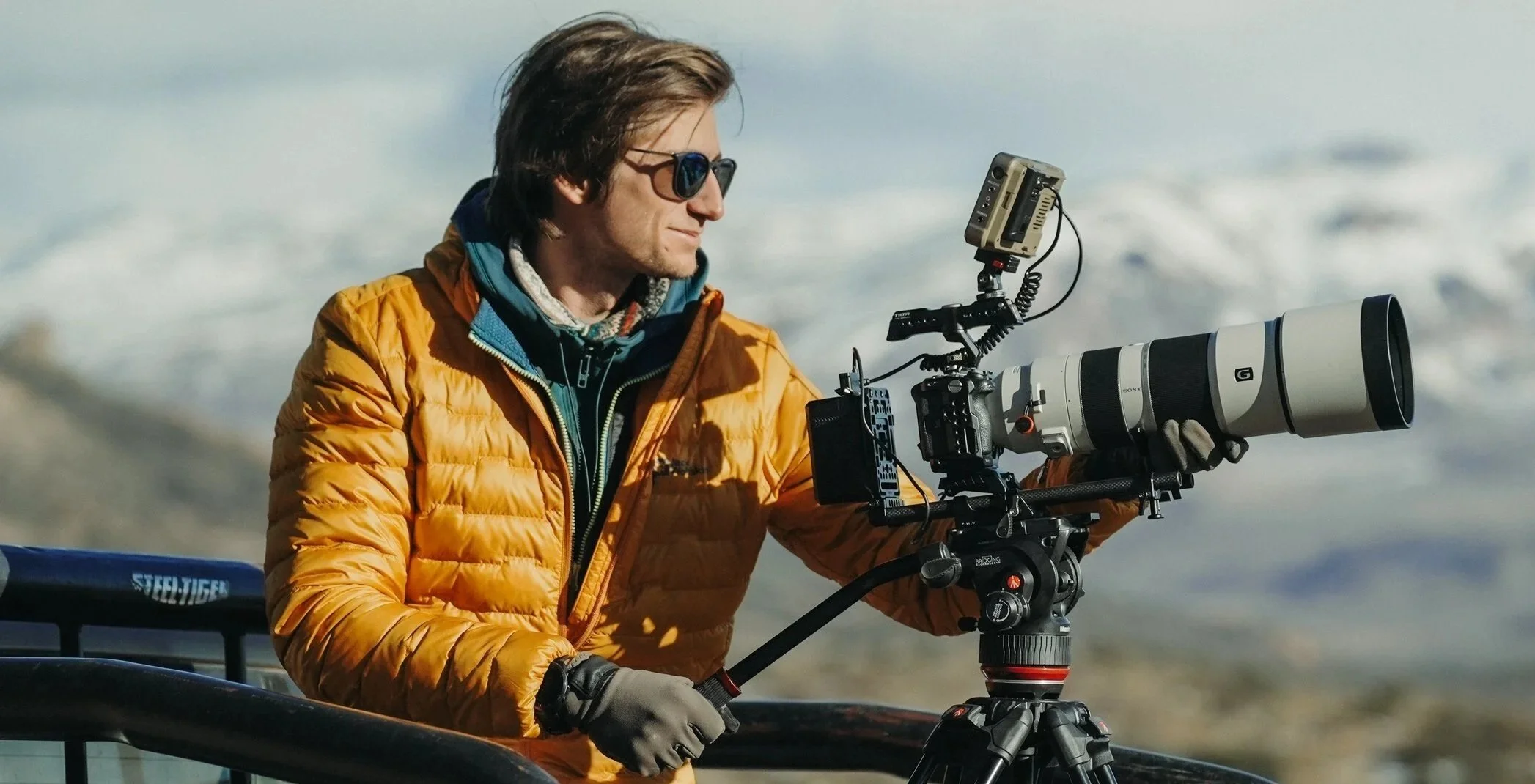 A person in a yellow jacket and sunglasses is operating a camera with a large telephoto lens outdoors, possibly in a snowy or mountainous area.