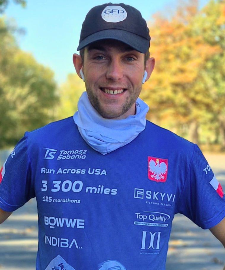 A man smiling in running gear outdoors during daytime with trees in the background. He is wearing a black cap, sports earphones, a blue running shirt with logos, and a neck gaiter.
