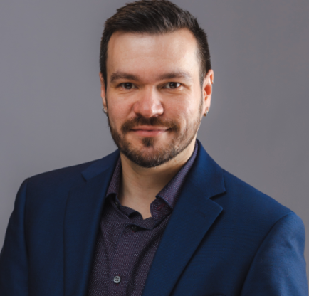 Headshot of a man with dark hair and a beard, wearing a navy suit and dark shirt, smiling against a gray background.