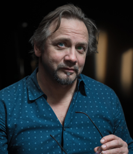 Portrait of a middle-aged man with light brown hair, a goatee, wearing a blue polka-dot shirt, holding a pair of glasses.