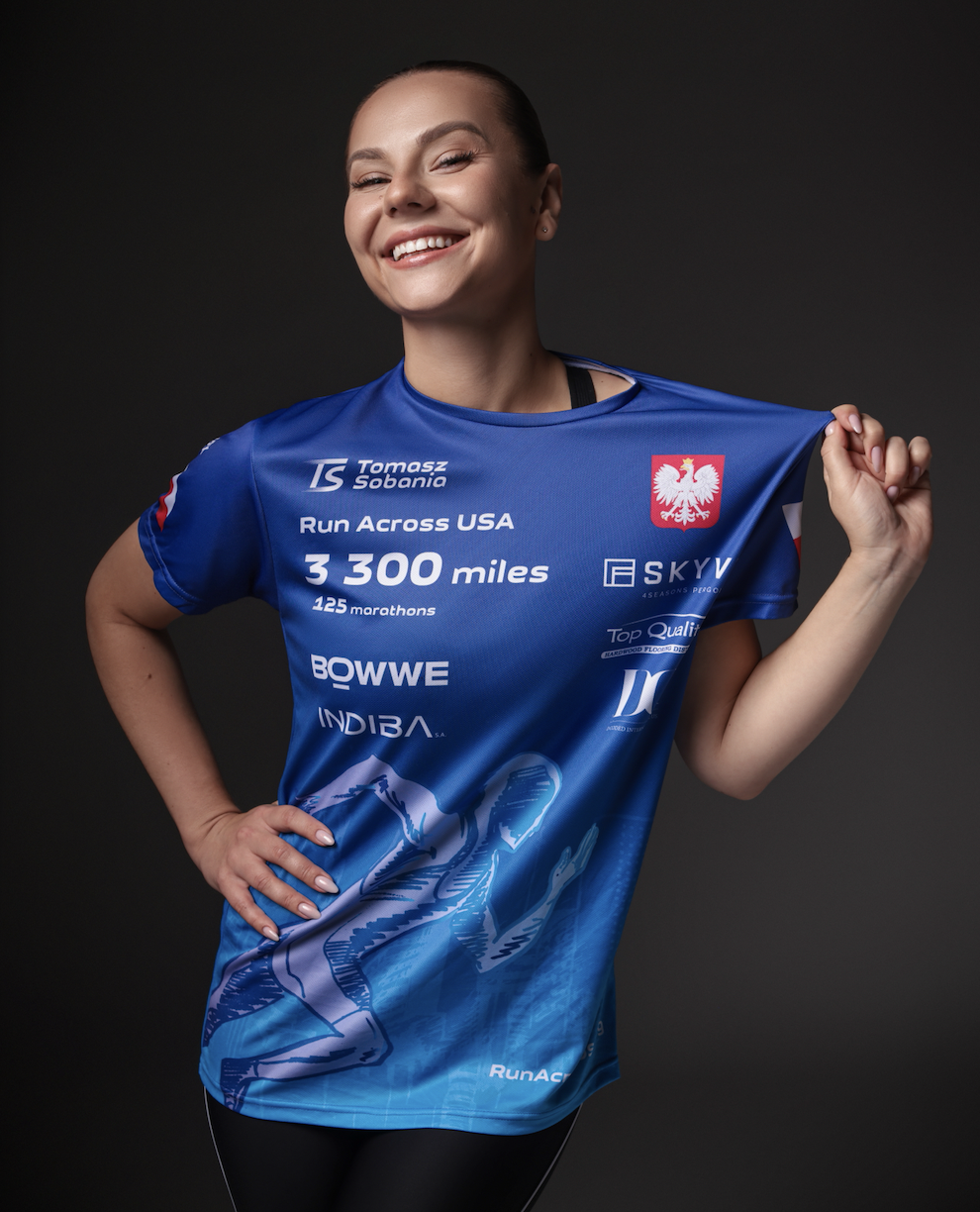 Polish pop singer DaggaNOW in a blue athletic shirt, holding up her shirt to reveal a logo and text related to running a 300-mile race across the USA.