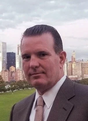 A man in a suit standing outdoors with a city skyline and green field in the background.