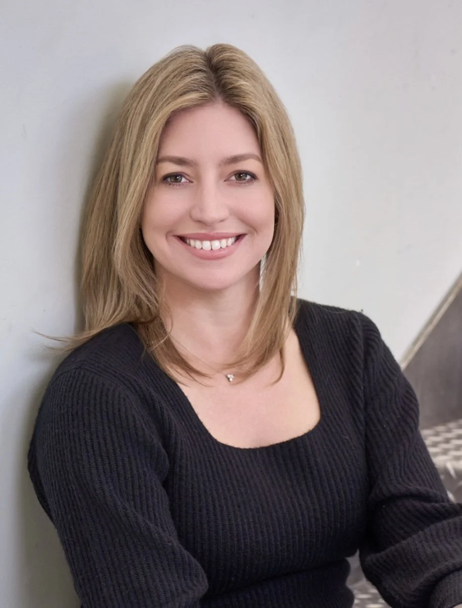 Courtney Hanna, lawyer turner executive producer/actress, shoulder-length blonde hair, smiling, wearing a black ribbed sweater, sitting against a light gray wall.