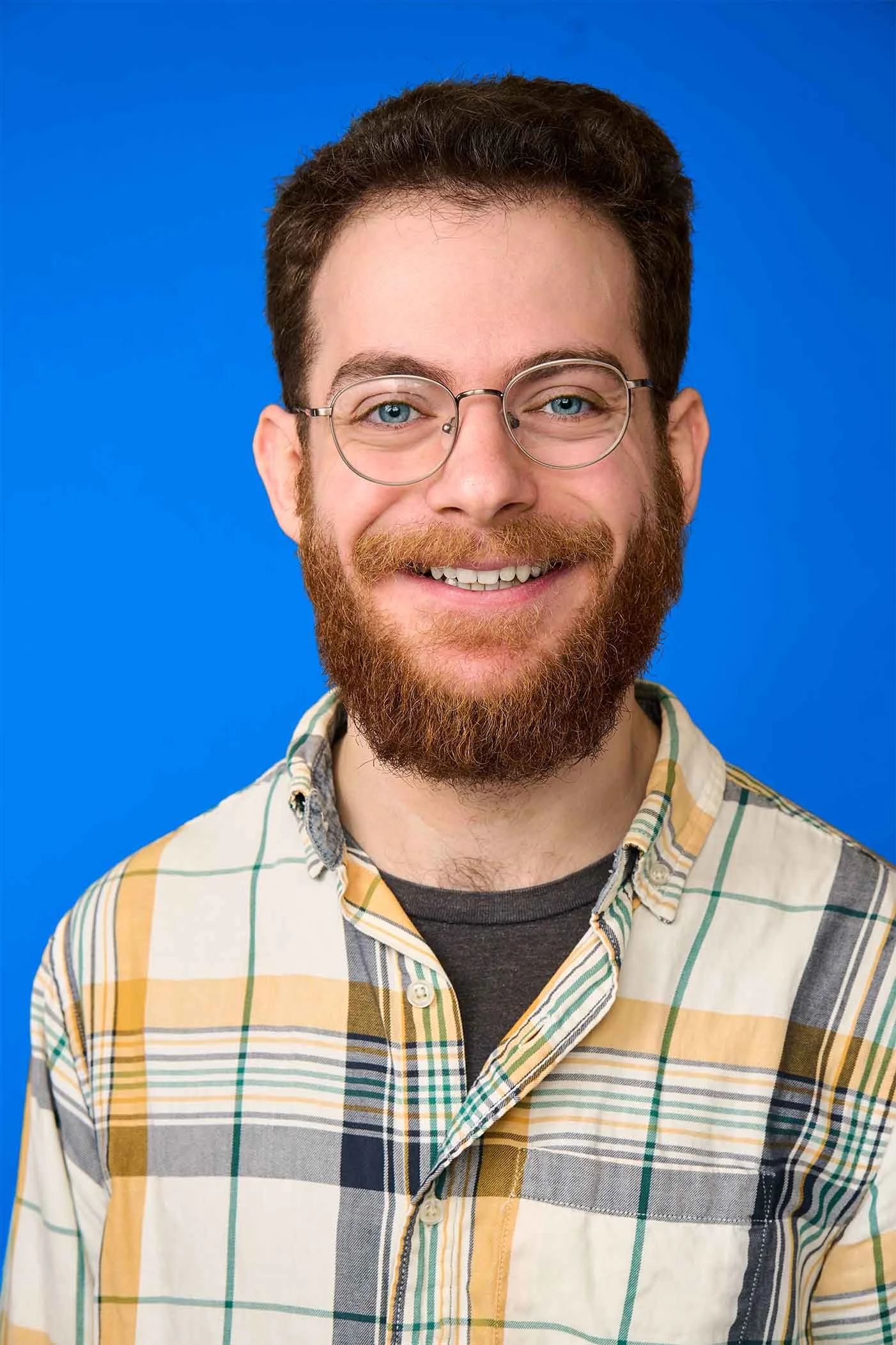Max Wexler, graphic designer/editor young man with glasses, a beard, and a mustache, smiling at the camera, wearing a plaid shirt, against a solid blue background.